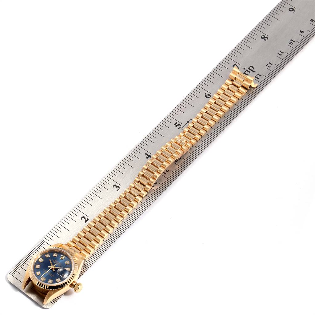 This image shows a top-down view of a Rolex President 69178 Women's Yellow Gold Blue Dial 69178 Women's Yellow Gold Blue Dial watch with a gold bracelet laid on a ruler.