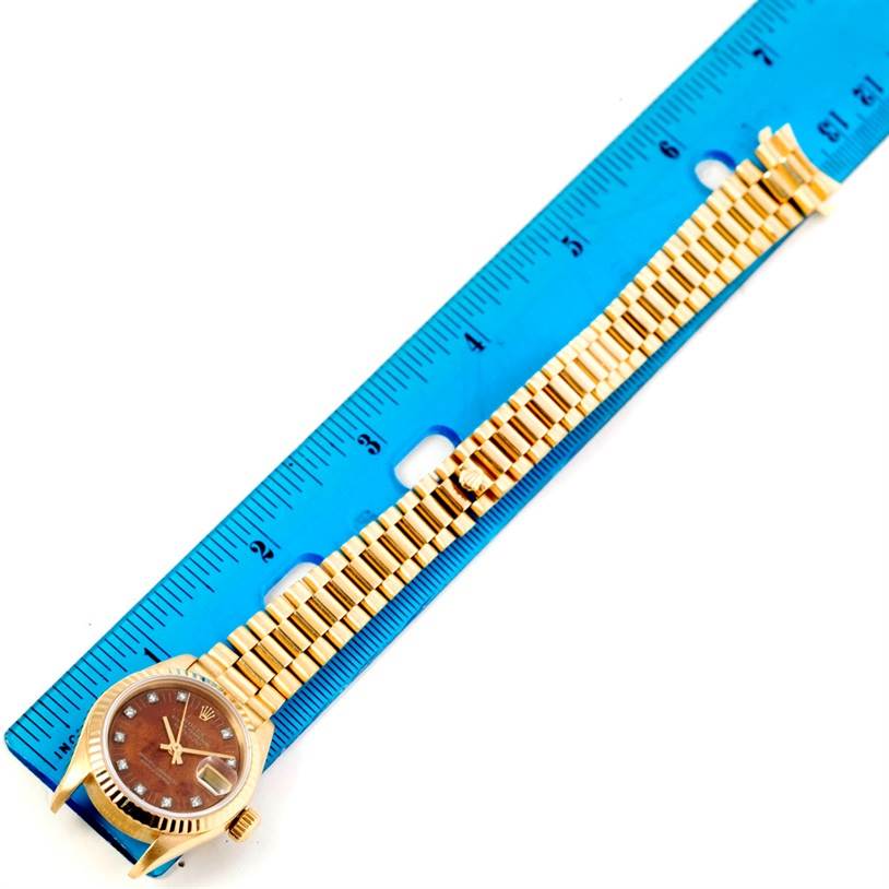 The image shows a Rolex President 69178 Women
s Yellow Gold Brown Dial 69178 Women
s Yellow Gold Brown Dial model watch placed alongside a blue measuring ruler with its face, bracelet, and crown visible.
