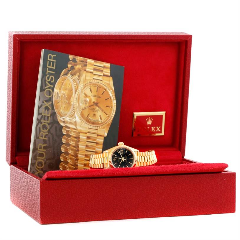 This image shows a frontal view of a Rolex President 69178 Women's Yellow Gold Black Dial 69178 Women's Yellow Gold Black Dial model watch inside a red case, including its booklet and logo.