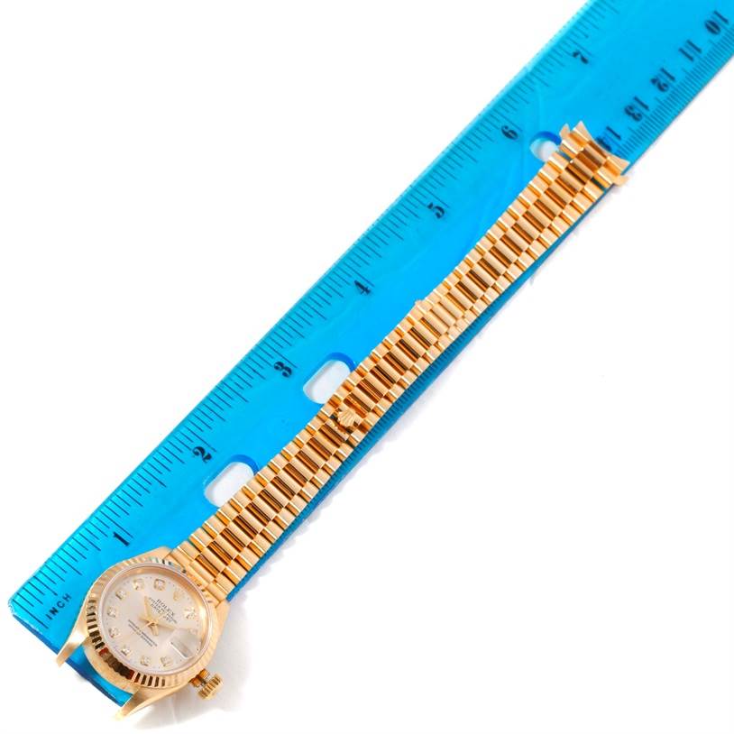 The image shows a top view of a Rolex President 69178 Women's Yellow Gold Silver Dial 69178 Women's Yellow Gold Silver Dial model watch with its golden bracelet aligned next to a blue ruler.