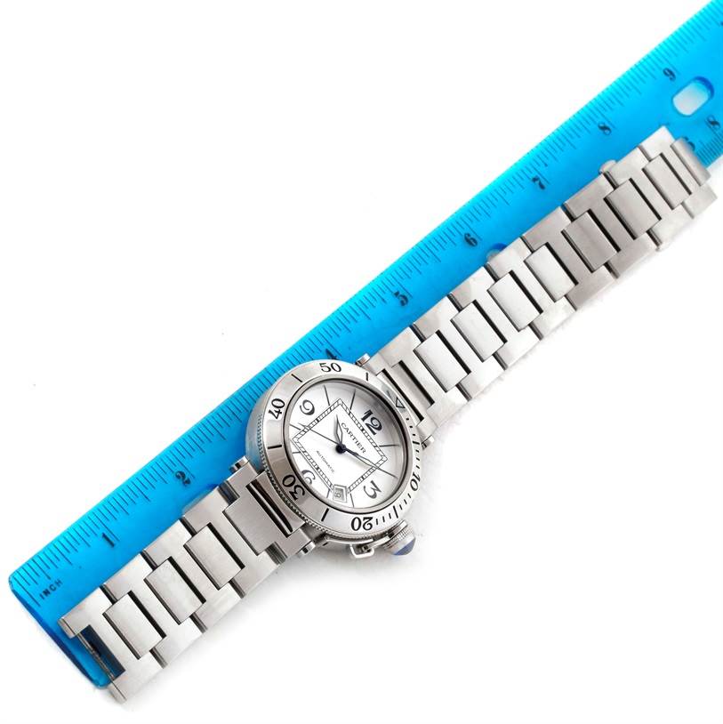 The image shows a Cartier Pasha W31080M7 Men's Stainless Steel Silver Dial W31080M7 Men's Stainless Steel Silver Dial watch placed diagonally with its bracelet extended and slightly wrapped around a blue ruler.