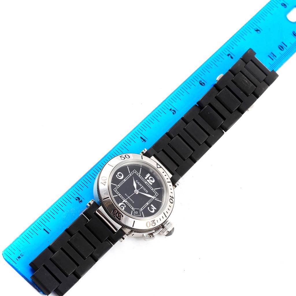 This image shows a top-down view of a Cartier Pasha W31077U2 Men's Stainless Steel Black Dial W31077U2 Men's Stainless Steel Black Dial watch with its black strap alongside a blue ruler for measurement.