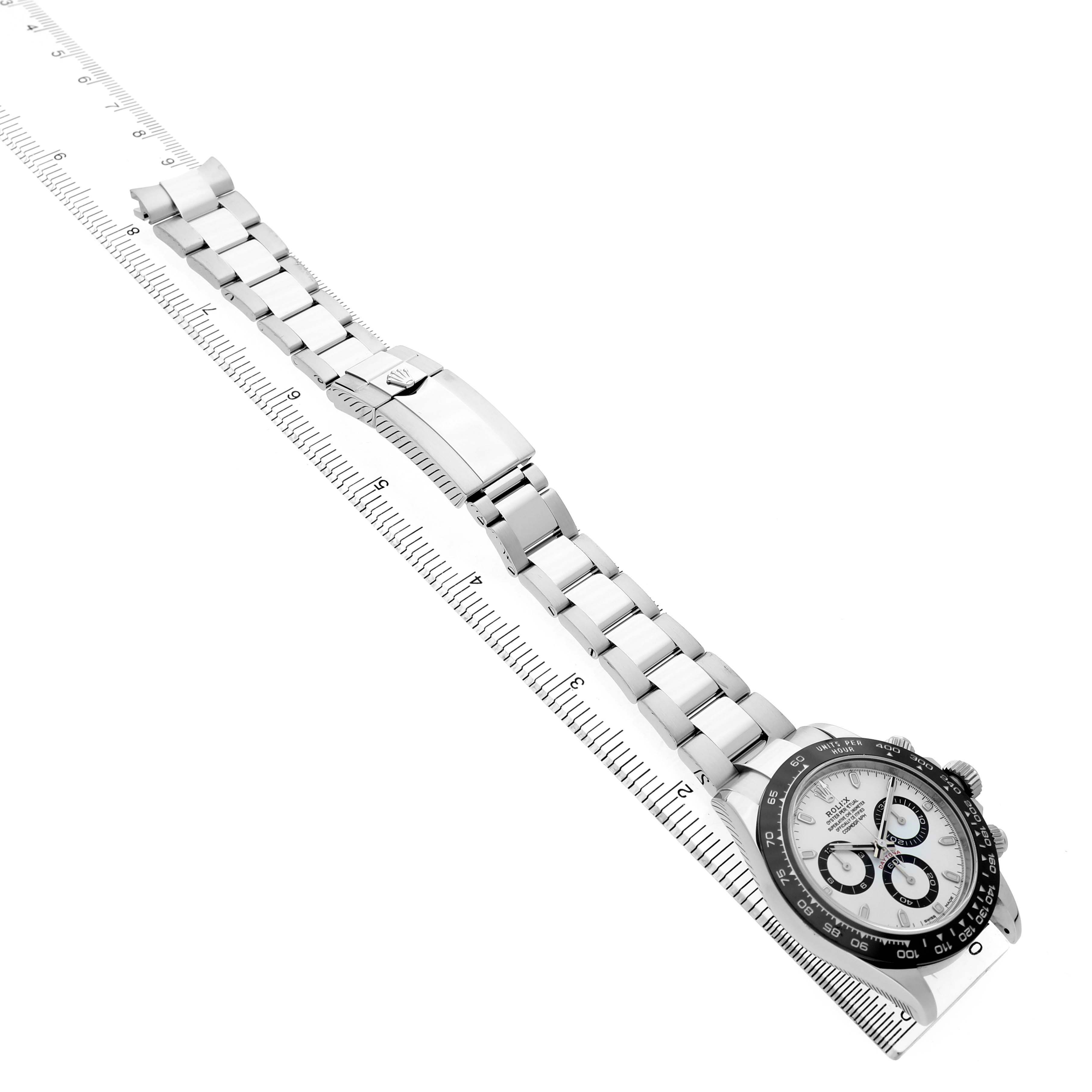 The Rolex Daytona 116500LN Men's Stainless Steel White Dial watch is shown from a side angle, displaying its bracelet, clasp, and dial with a measuring ruler beside it.