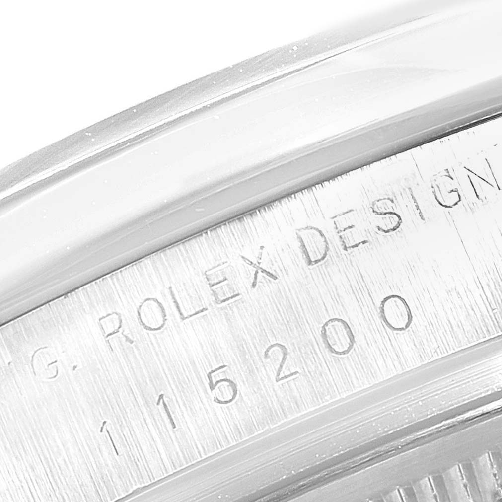 The image shows an angled close-up of the engraved serial number on the case of a Rolex Date 115200 Men's Stainless Steel Silver Dial 115200 Men's Stainless Steel Silver Dial model watch.