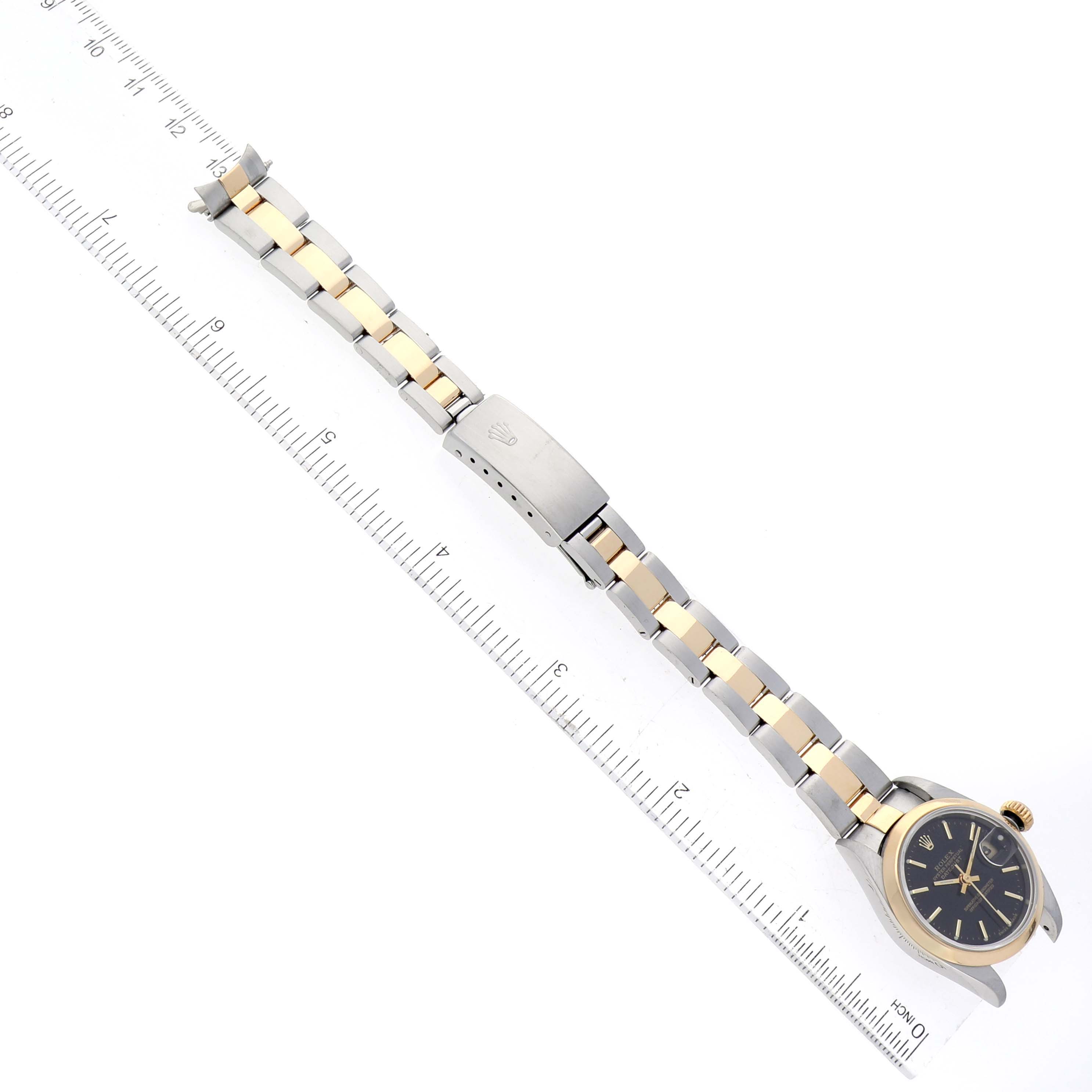The image shows a Rolex Datejust 79163 Women's Steel and Gold (two tone) Black Dial 79163 Women's Steel and Gold (two tone) Black Dial watch from an aerial view, featuring its face, two-tone metal strap, and clasp on a measuring ruler.