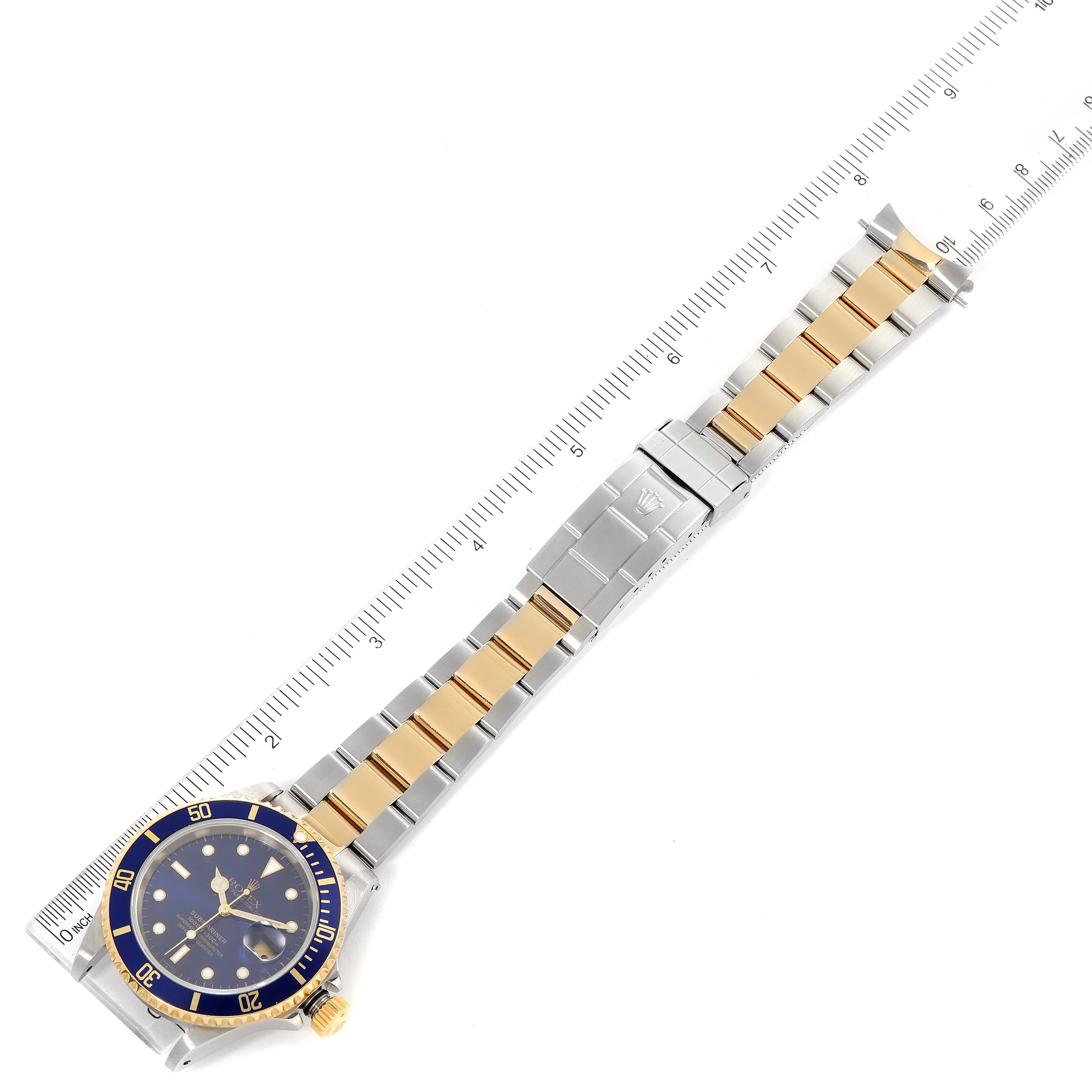 The image shows a Rolex Submariner 16613 Men's Steel and Gold (two tone) Blue Dial 16613 Men's Steel and Gold (two tone) Blue Dial watch placed horizontally with a ruler beside it, displaying the full bracelet and the watch face.
