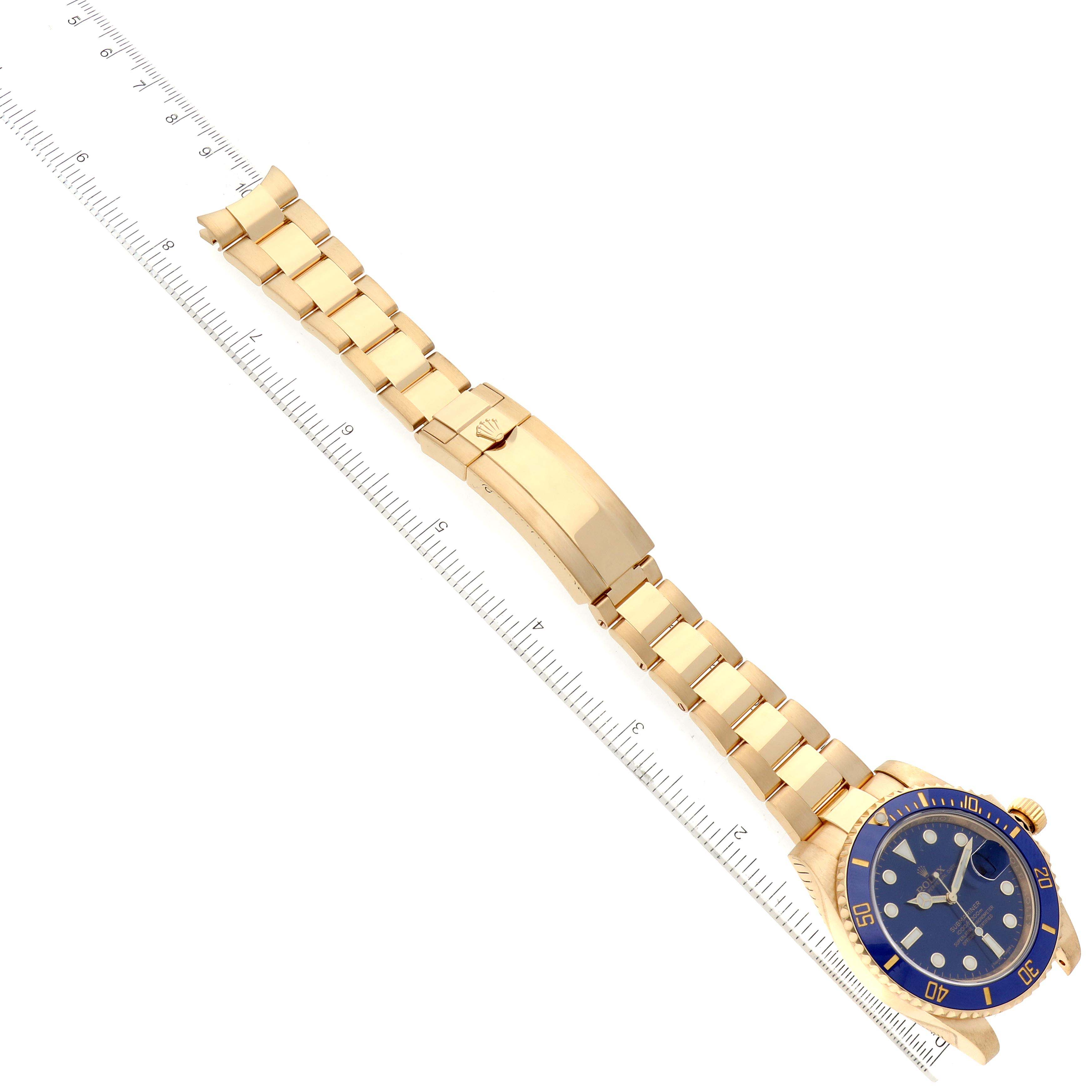 The Rolex Submariner 116618 Men's Yellow Gold Blue Dial 116618 Men's Yellow Gold Blue Dial watch is shown at an angled top-down view, displaying its gold bracelet, blue dial, bezel, and clasp near a ruler.