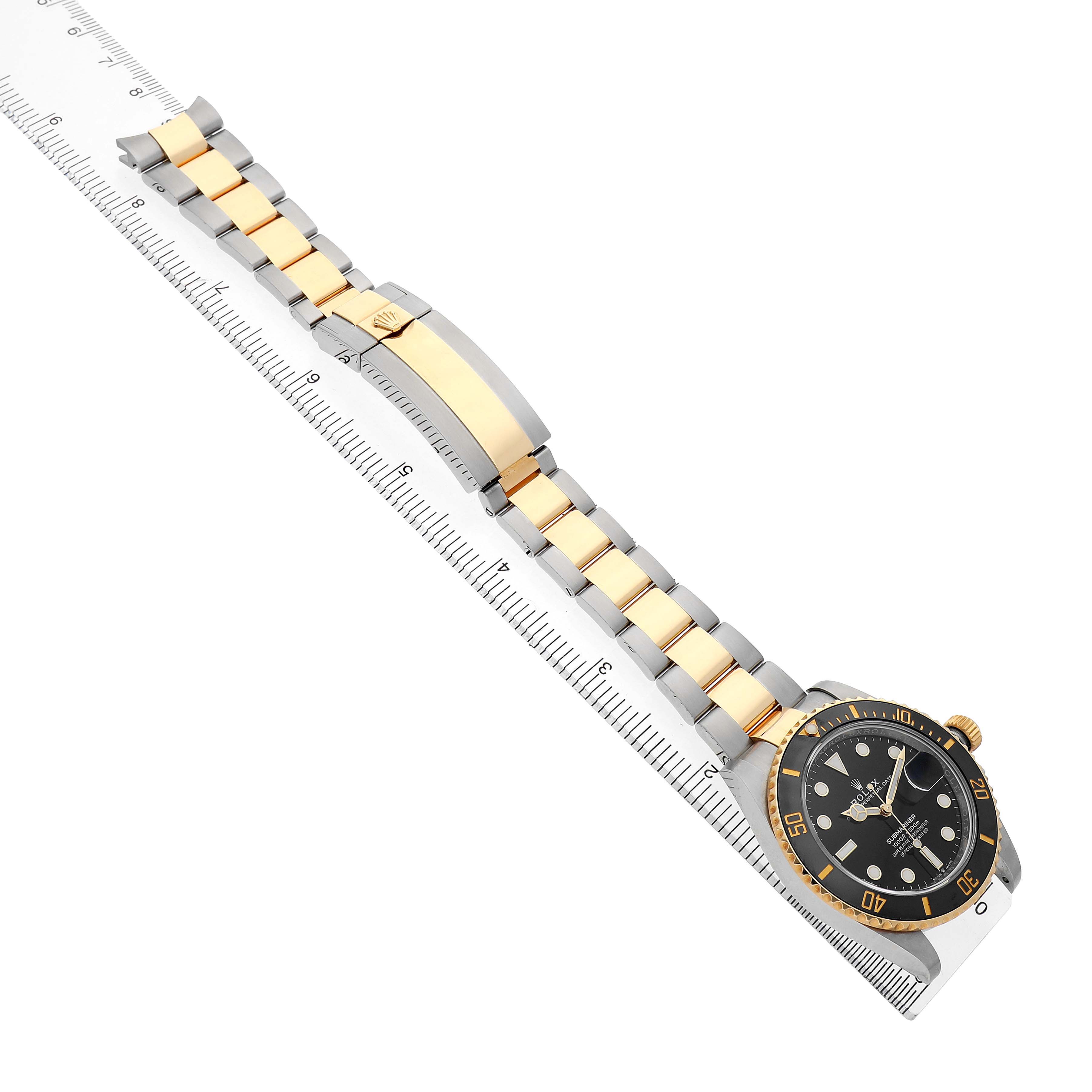 The Rolex Submariner M126613LN-0002 Men's Steel and Gold (two tone) Black Dial watch is shown at an angle displaying the face, bezel, crown, and full bracelet on a ruler.