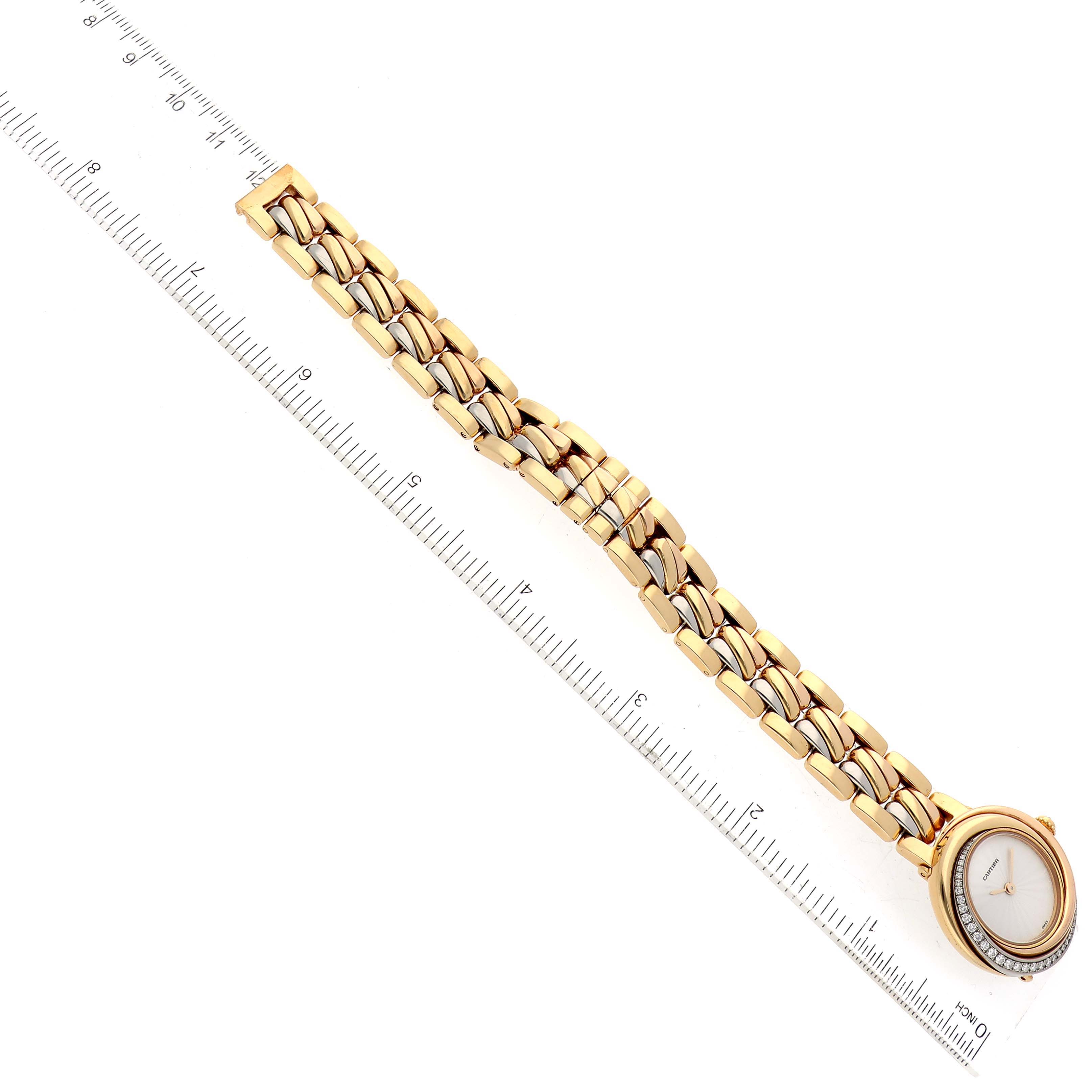 The image shows a Cartier Trinity WG200151 Women's Yellow Gold Silver Dial WG200151 Women's Yellow Gold Silver Dial model watch laid flat next to a ruler, highlighting its gold bracelet and oval face.