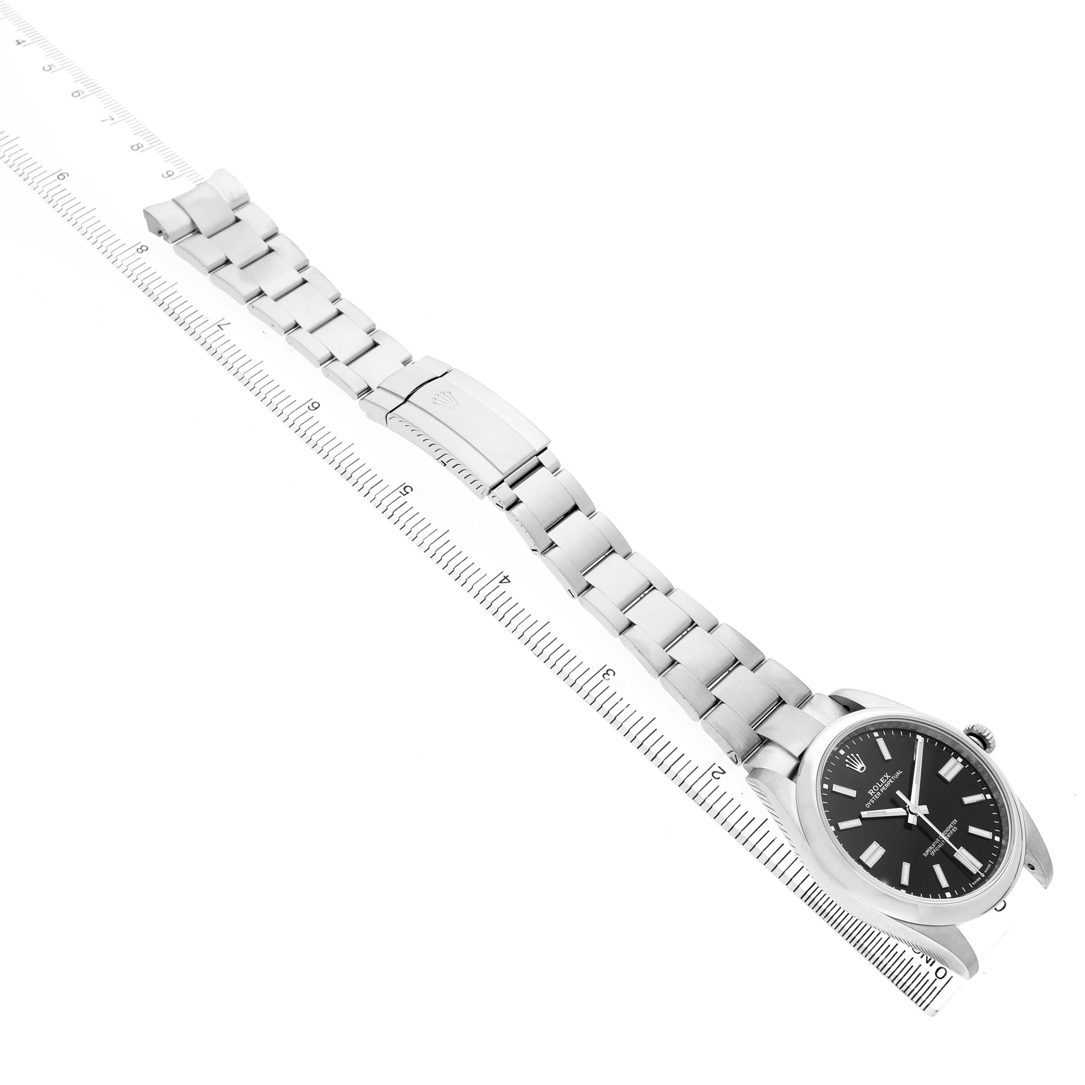 The image shows a Rolex Oyster Perpetual M124300-0002 Men's Stainless Steel Black Dial watch at an angled top view, displaying the face, case, and full bracelet next to a ruler.