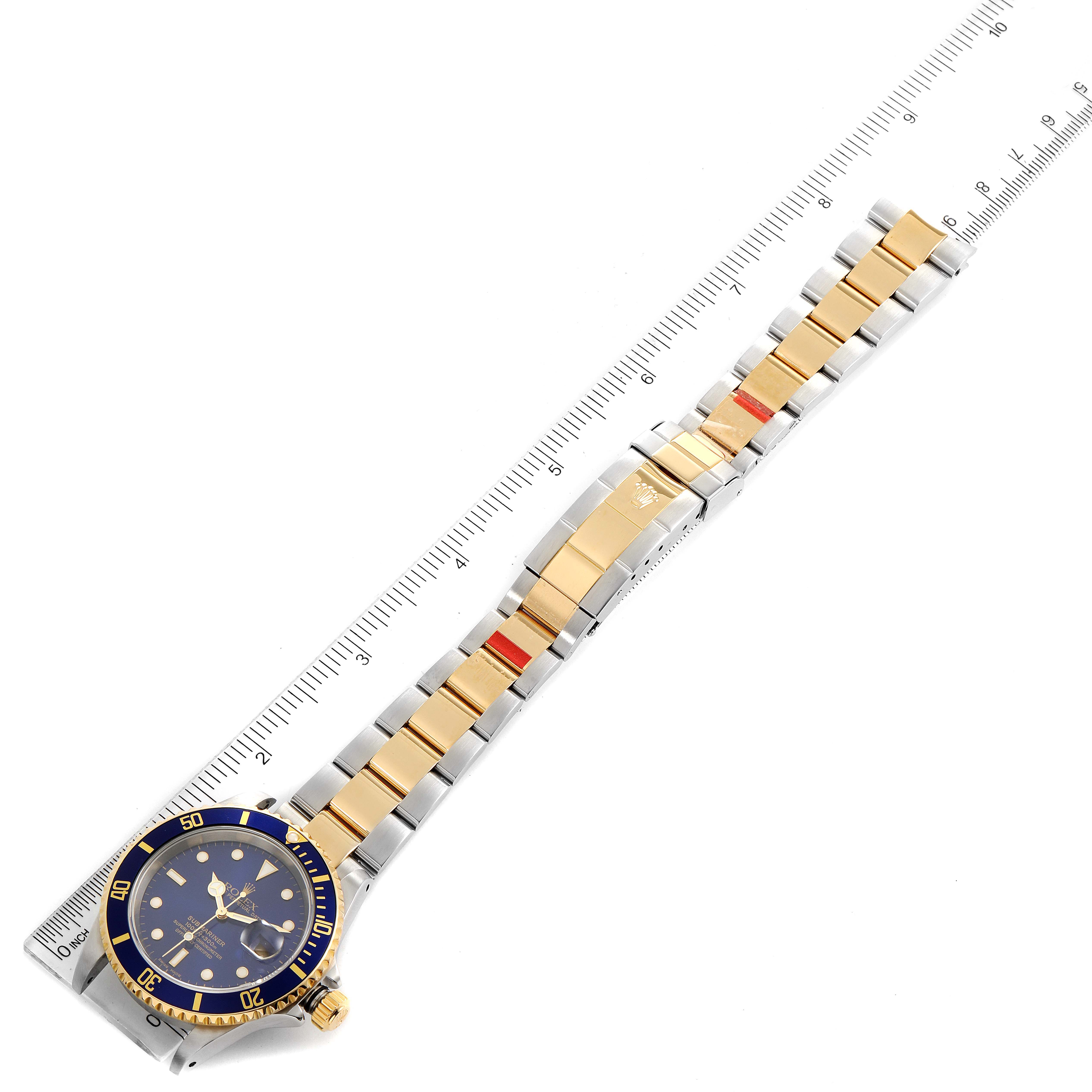 The image shows a Rolex Submariner 16613 Men
s Steel and Gold (two tone) Blue Dial 16613 Men
s Steel and Gold (two tone) Blue Dial watch laid flat next to a ruler, highlighting its gold and silver bracelet and blue dial.