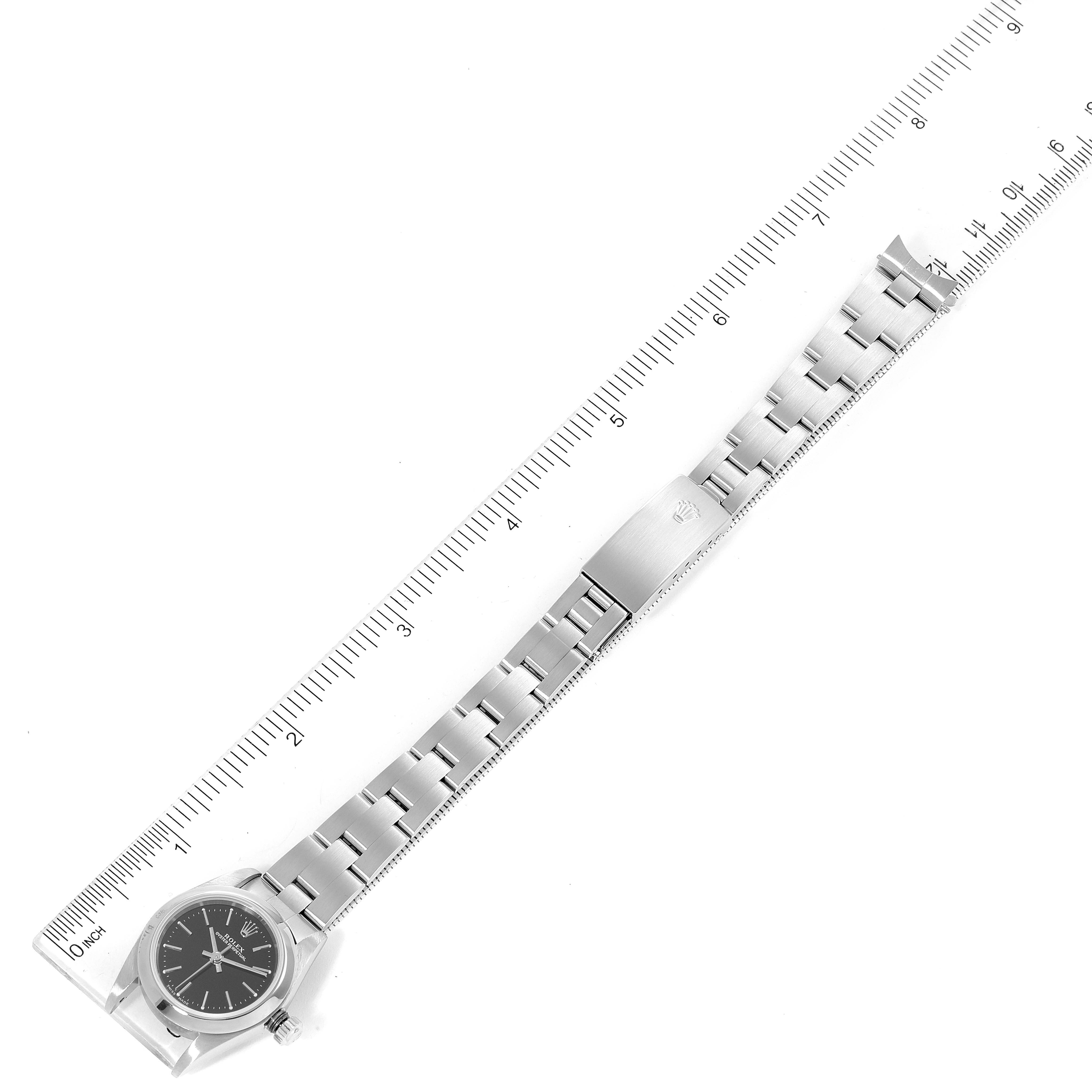 The image shows a top view of a Rolex Oyster Perpetual 76080 Women's Stainless Steel Black Dial 76080 Women's Stainless Steel Black Dial watch with its full metal bracelet extended alongside a ruler.