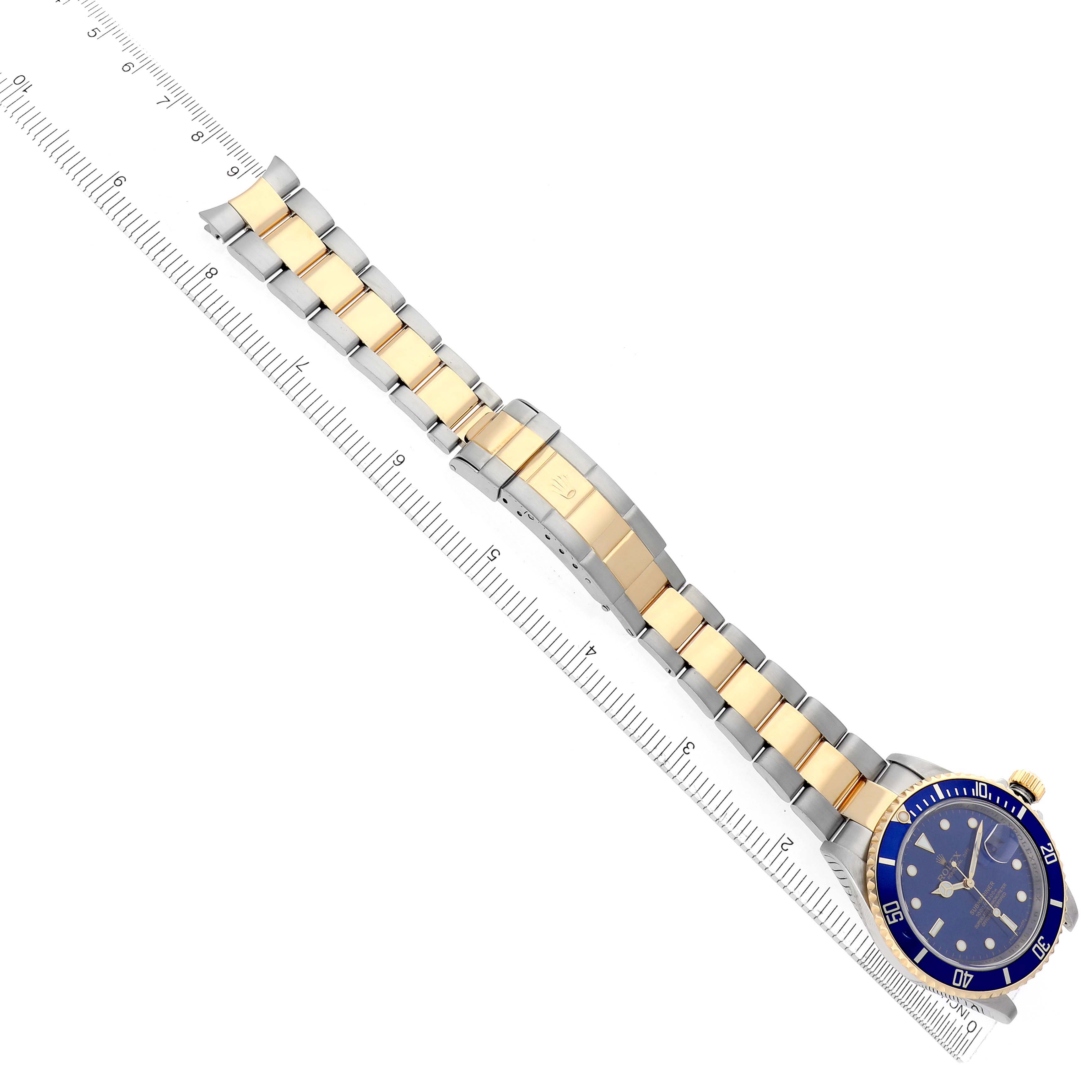 The image shows a Rolex Submariner 16613 Men
s Steel and Gold (two tone) Blue Dial 16613 Men
s Steel and Gold (two tone) Blue Dial at a diagonal angle, highlighting its blue dial, bezel, and two-tone bracelet across a ruler.