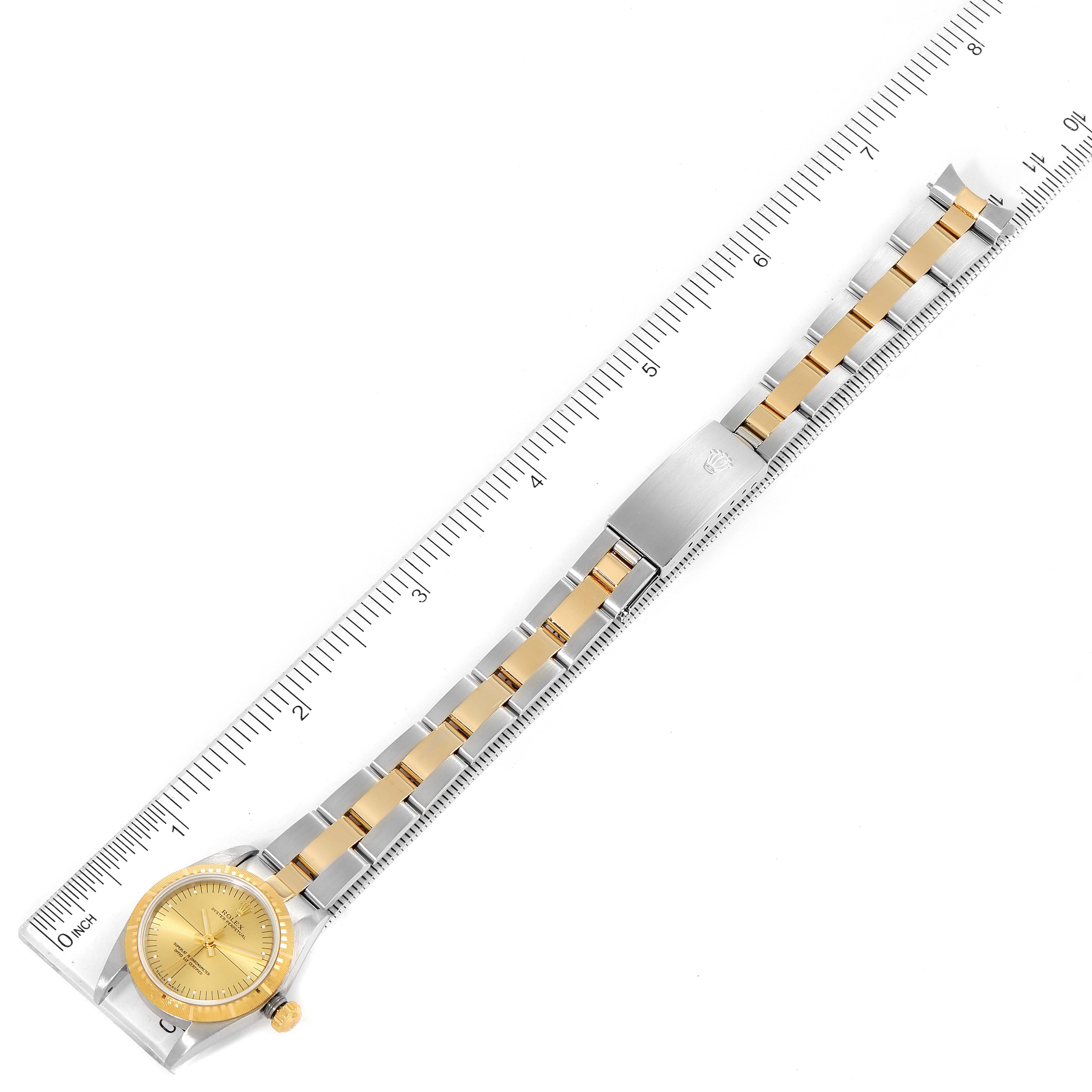 The image shows a Rolex Oyster Perpetual 76243 Women
s Steel and Gold (two tone) Champagne Dial 76243 Women
s Steel and Gold (two tone) Champagne Dial watch laid flat with a metal bracelet and a two-tone (gold and silver) band, alongside a ruler.