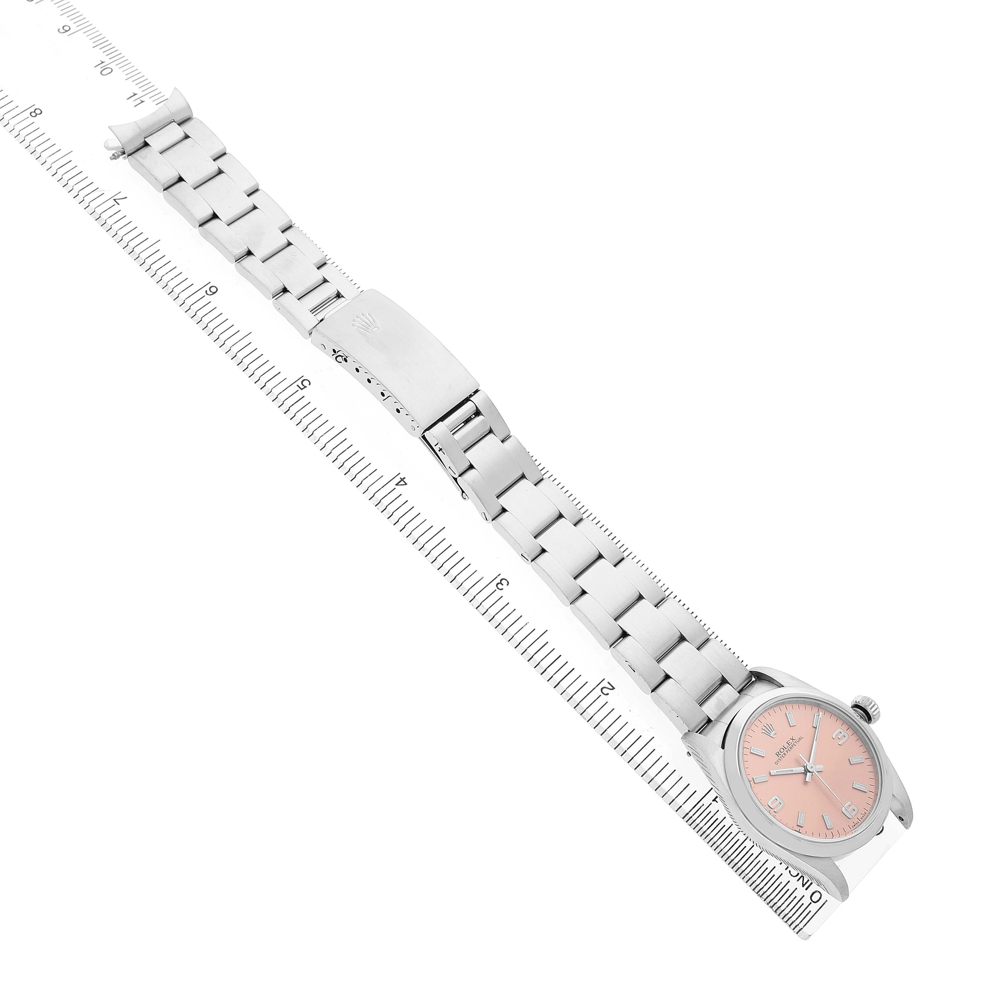 The Rolex Oyster Perpetual 77200 Women's Stainless Steel Pink Dial watch is displayed at an angle, showing the face, bracelet, and clasp alongside a ruler for scale.