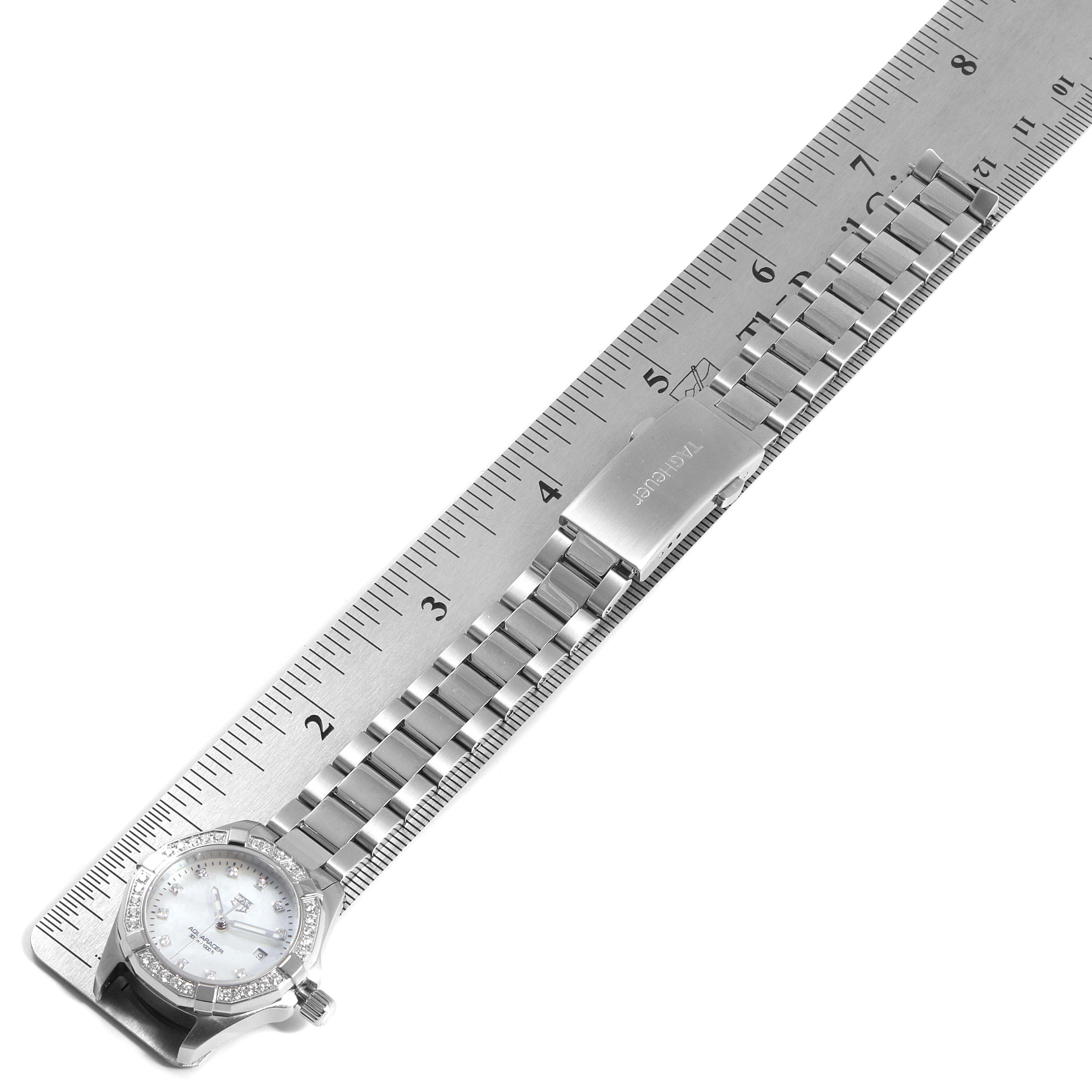 The image shows the Tag Heuer Aquaracer WBD1415.BA0741 Women's Stainless Steel Mother of Pearl Dial WBD1415.BA0741 Women's Stainless Steel Mother of Pearl Dial watch with a metal strap, positioned diagonally on a metal ruler.