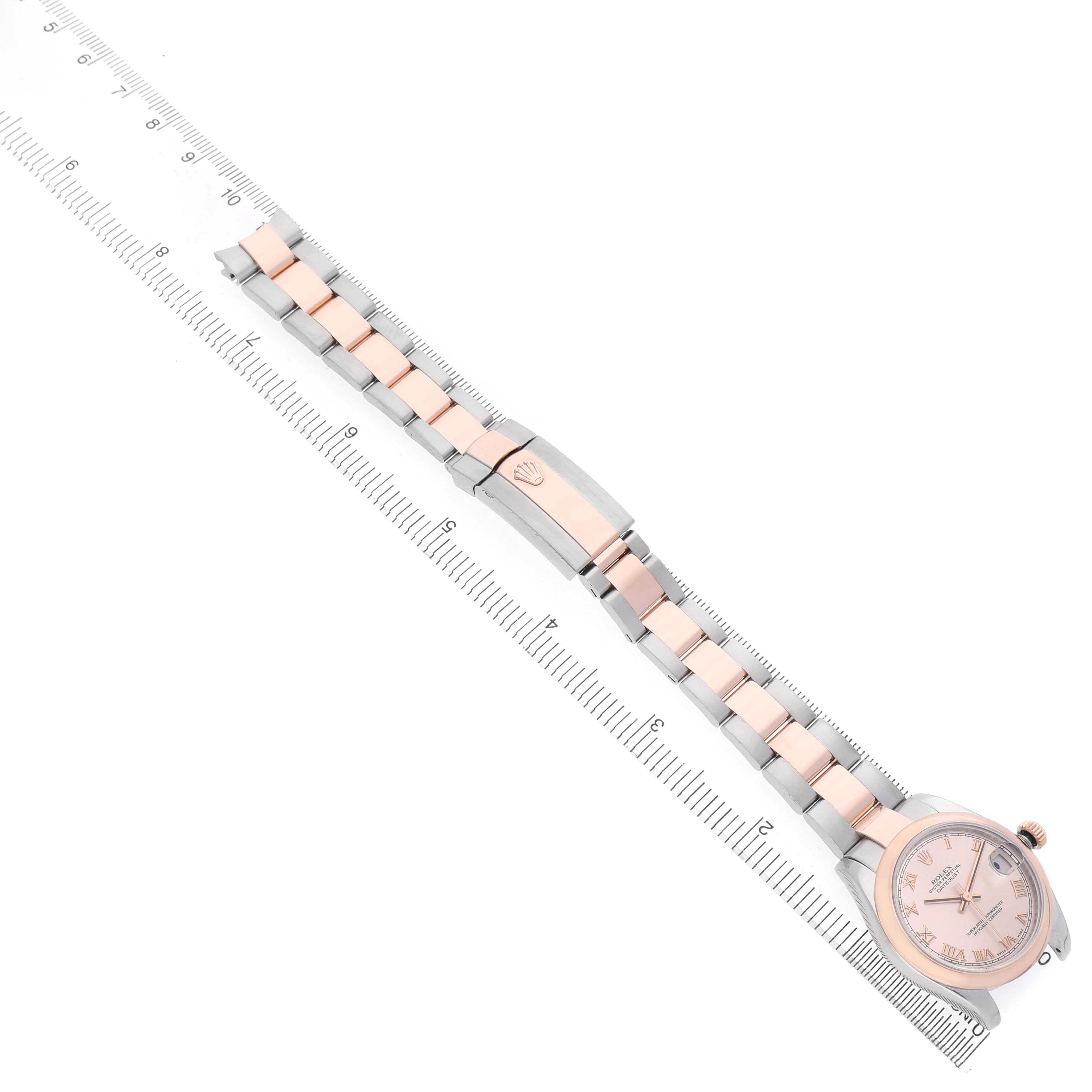 The Rolex Mid-Size 178241 Women's Steel and Gold (two tone) Pink Dial watch is shown from an aerial view, highlighting the face, bracelet, and clasp next to a ruler for scale.