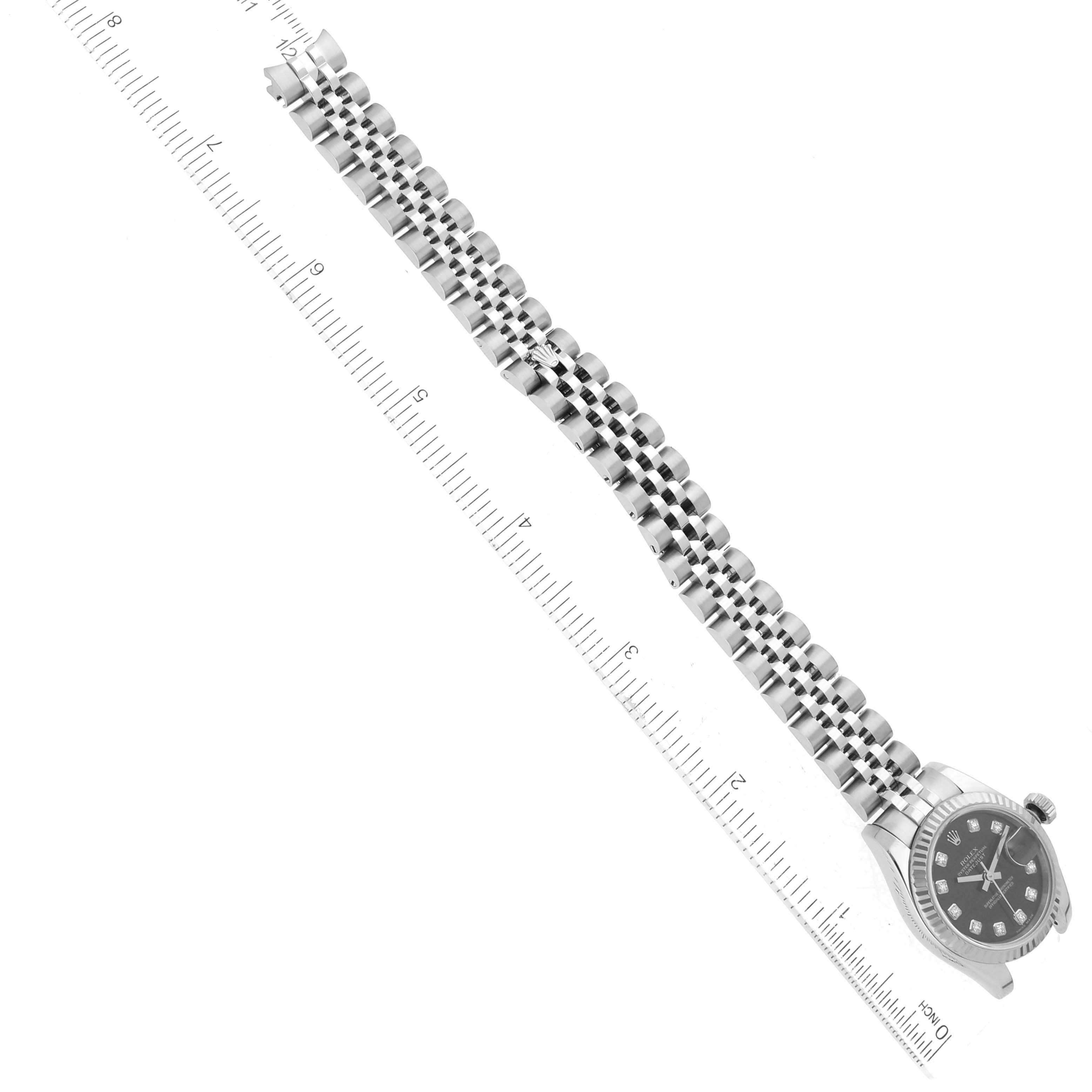 The image shows a diagonal top view of a Rolex Datejust 179174 Women
s Stainless Steel Black Dial 179174 Women
s Stainless Steel Black Dial watch with a metal bracelet placed against a ruled background.