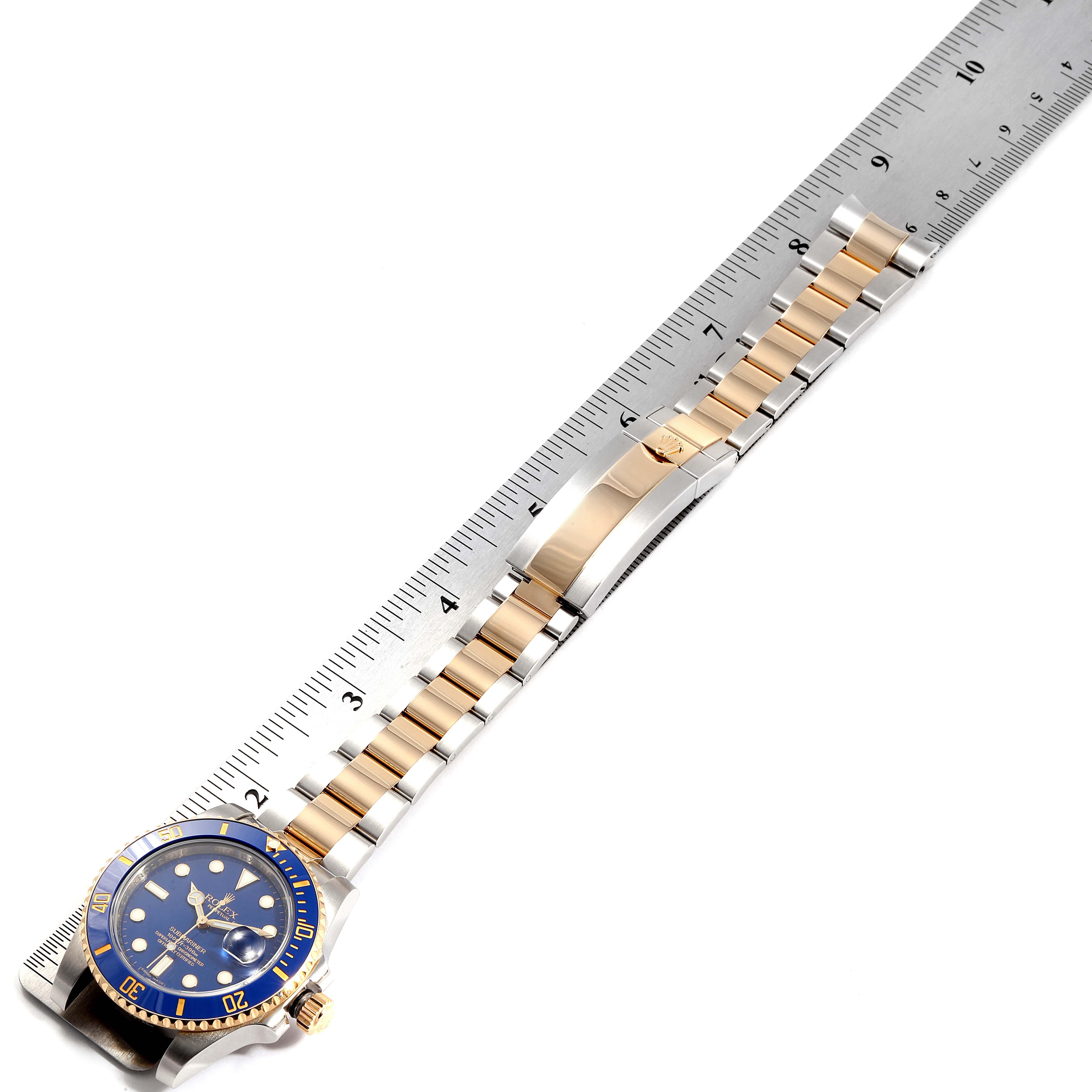 The image shows a Rolex Submariner 116613 Men
s Steel and Gold (two tone) Blue Dial 116613 Men
s Steel and Gold (two tone) Blue Dial watch with a blue dial and a two-tone metal bracelet laid next to a ruler.