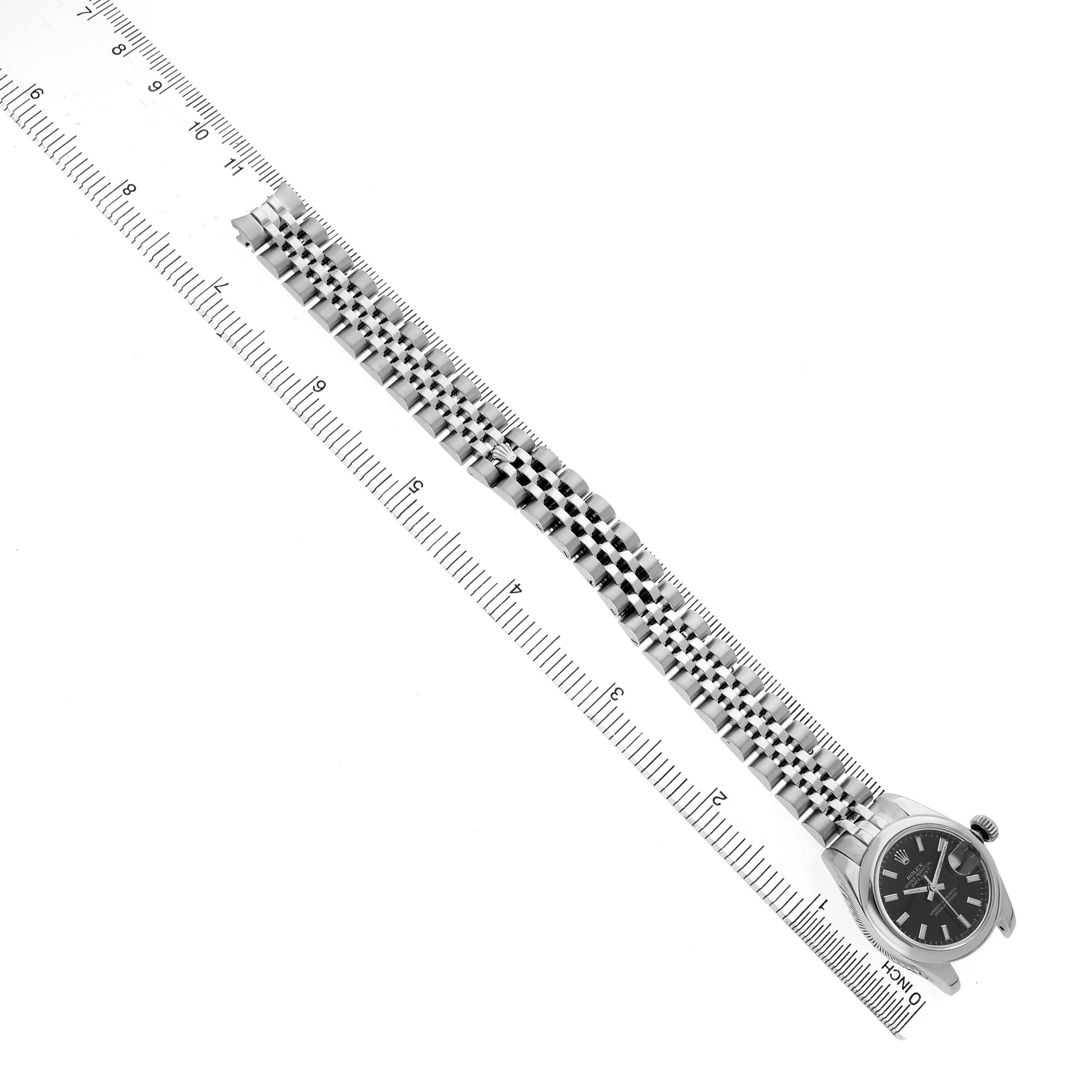 The Rolex Datejust 179160 Women's Stainless Steel Black Dial watch is shown from a top-down angle with its full bracelet and case, alongside a ruler for scale.