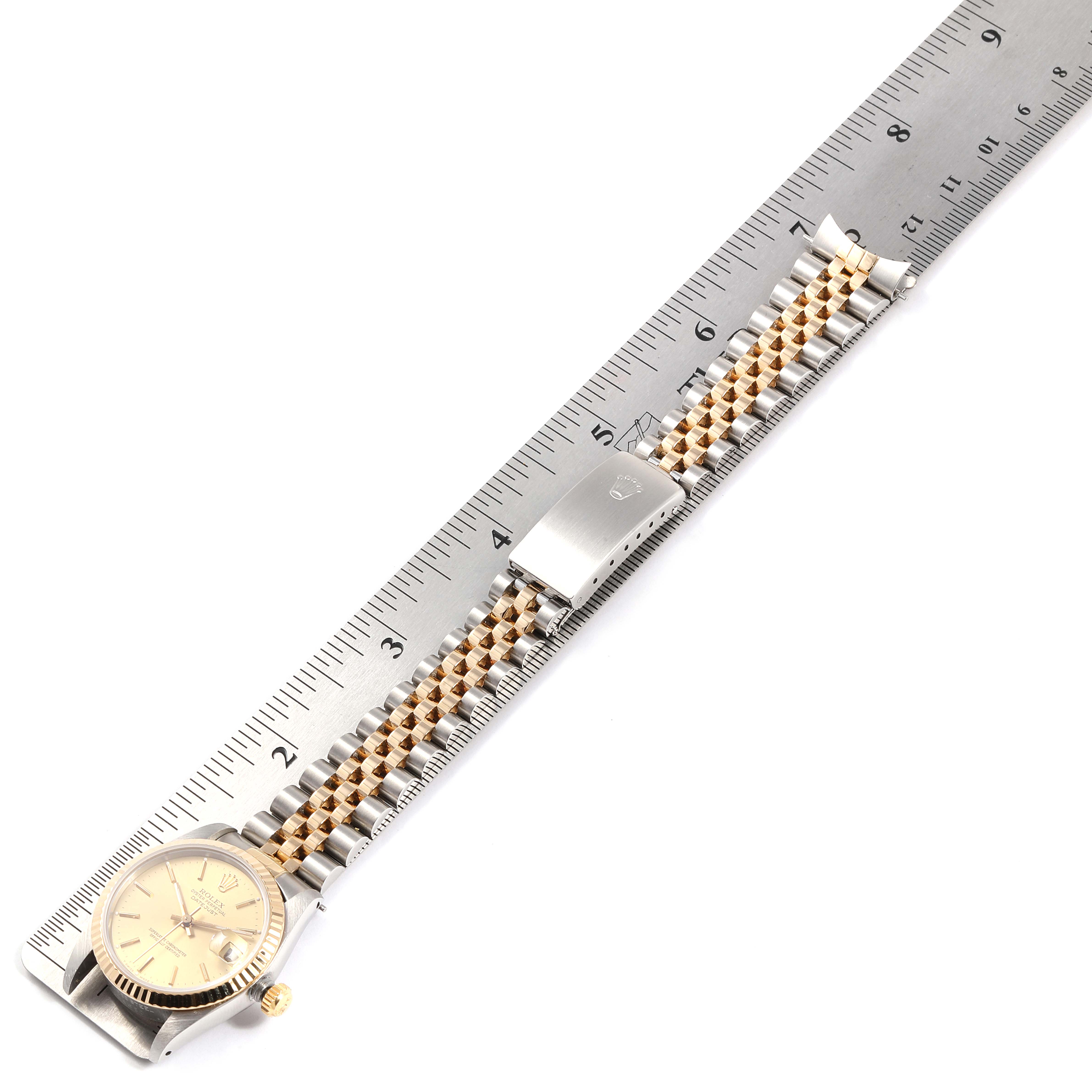 The image shows a Mid-Size Rolex watch lying flat next to a ruler, displaying its dial, crown, and metal bracelet with a clasp.