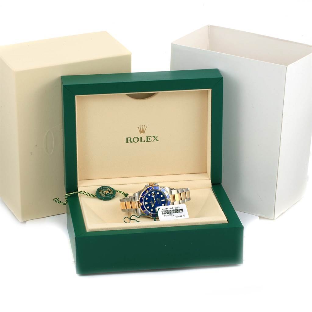The Rolex Submariner 116613 Men
s Steel and Gold (two tone) Blue Dial 116613 Men
s Steel and Gold (two tone) Blue Dial watch is displayed in its open green and cream-colored box, with packaging and tags visible.