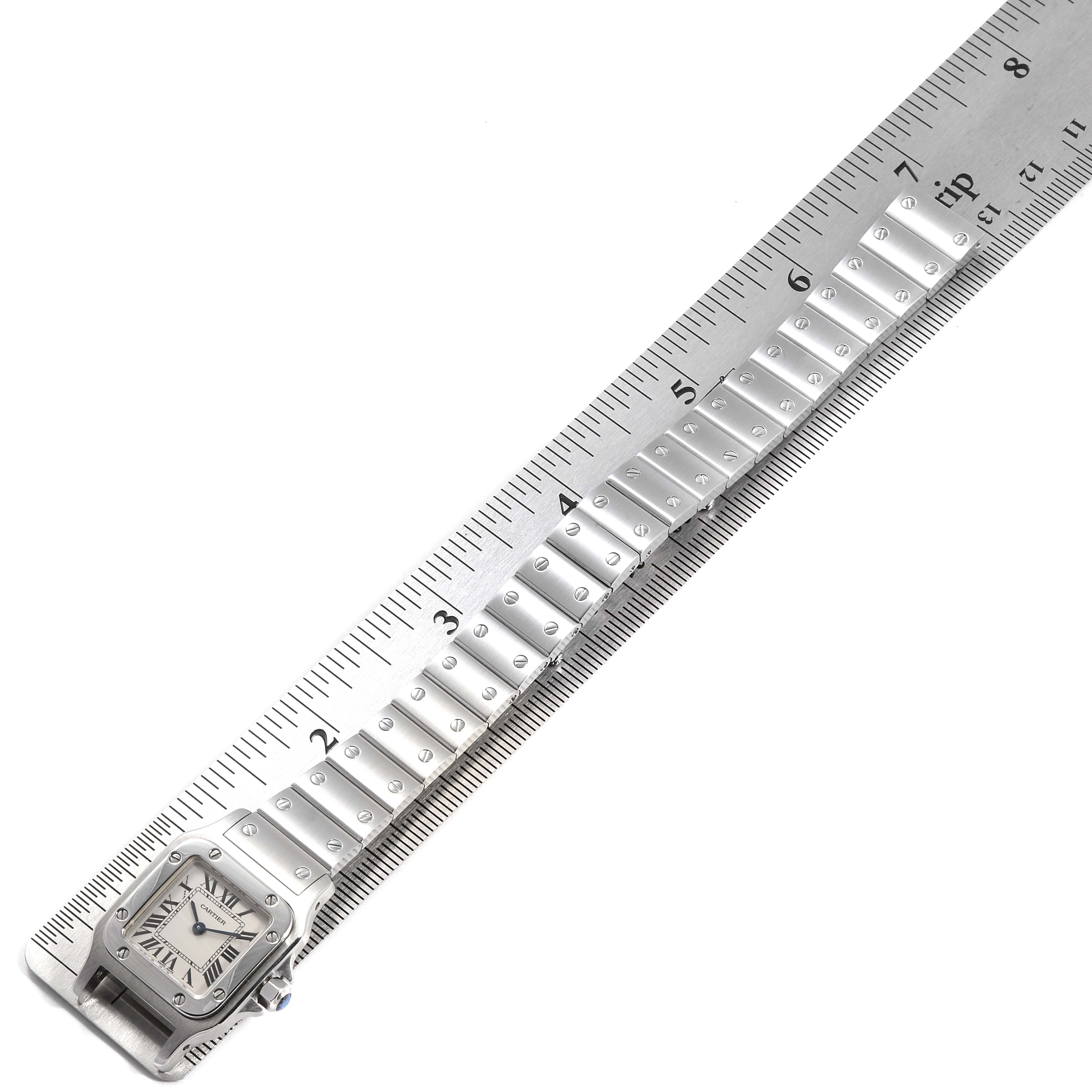The image shows a Cartier Santos W20056D6 Women's Stainless Steel Silver Dial W20056D6 Women's Stainless Steel Silver Dial watch laid flat against a ruler, displaying its face and part of the metal bracelet.