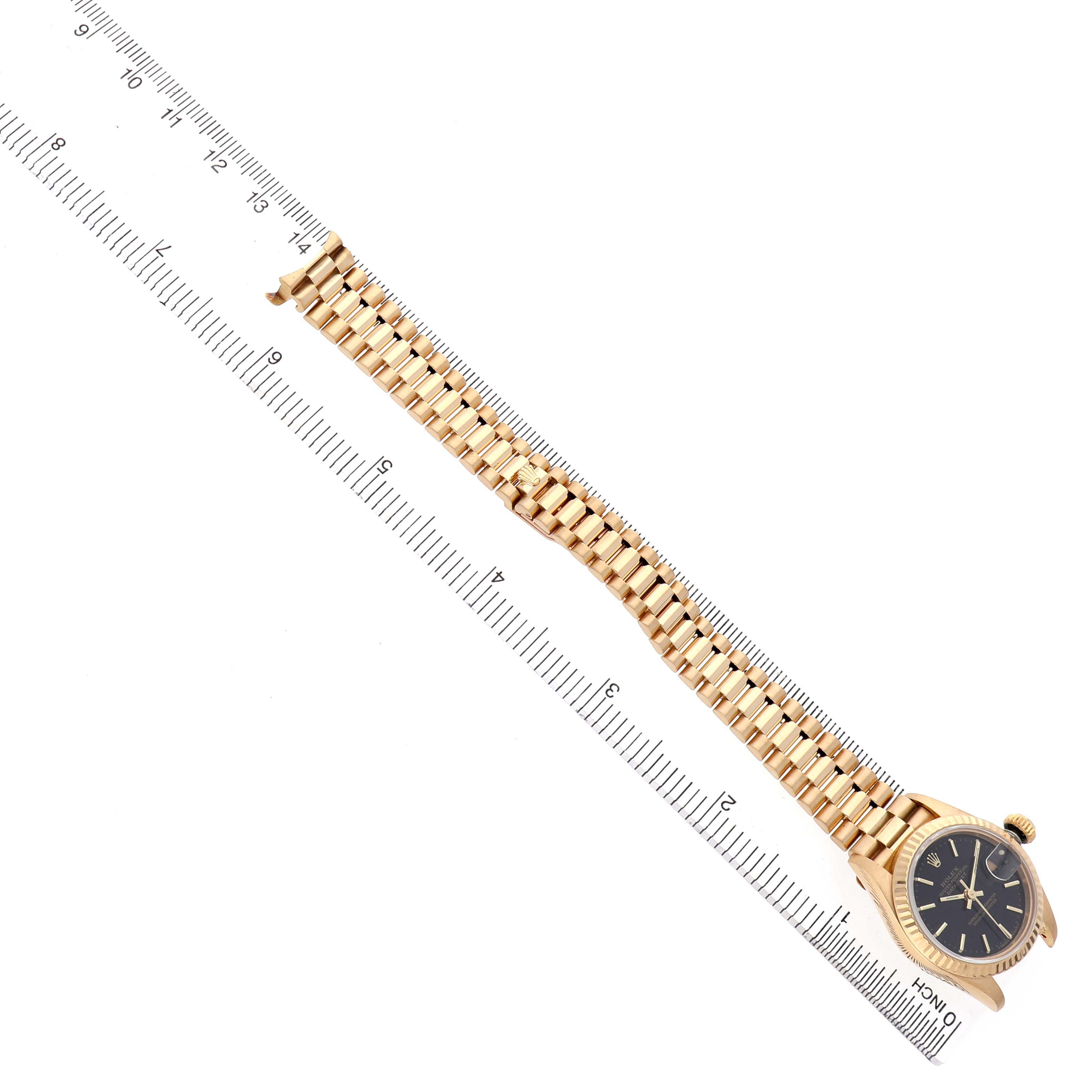 The Rolex President 69178 Women's Yellow Gold Black Dial model watch is shown from a top-down angle, displaying the full bracelet and face alongside a ruler for scale.