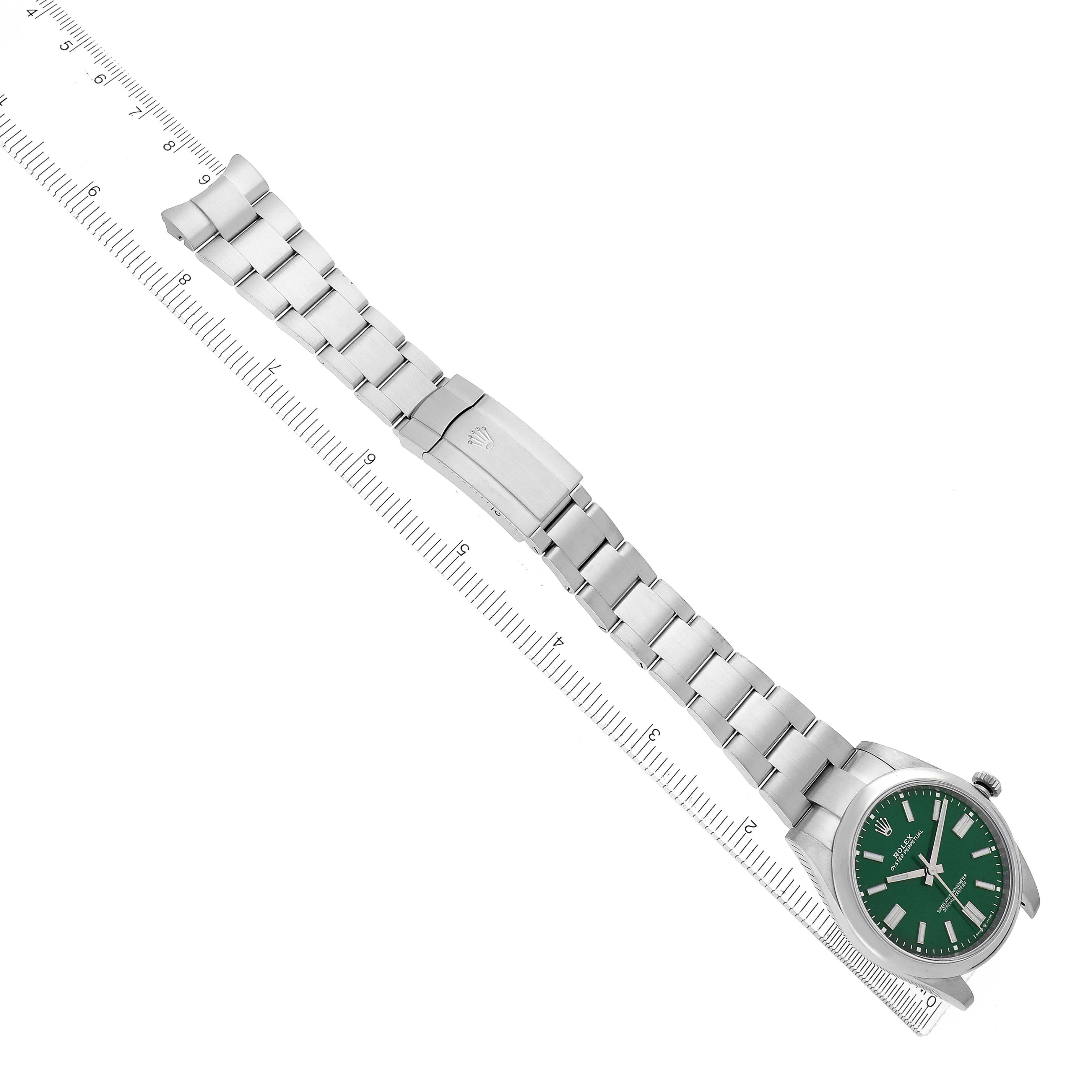The Rolex Oyster Perpetual model 124300 is shown from an overhead angle, highlighting the watch case, green dial, and entire bracelet laid out alongside a ruler.