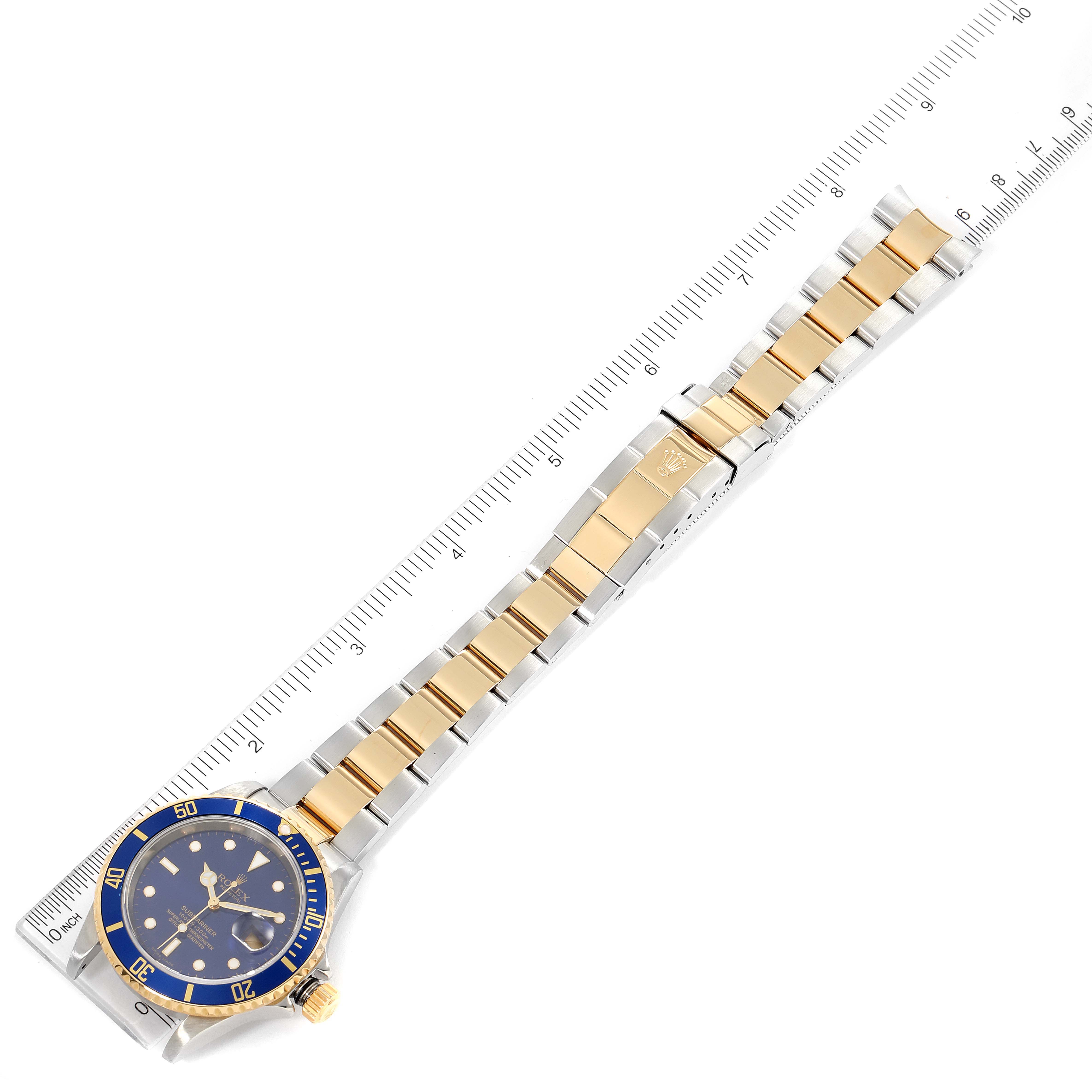 This image shows a Rolex Submariner 16613 Men's Steel and Gold (two tone) Blue Dial 16613 Men's Steel and Gold (two tone) Blue Dial watch laid flat beside a ruler, displaying the dial, bezel, bracelet, and clasp.
