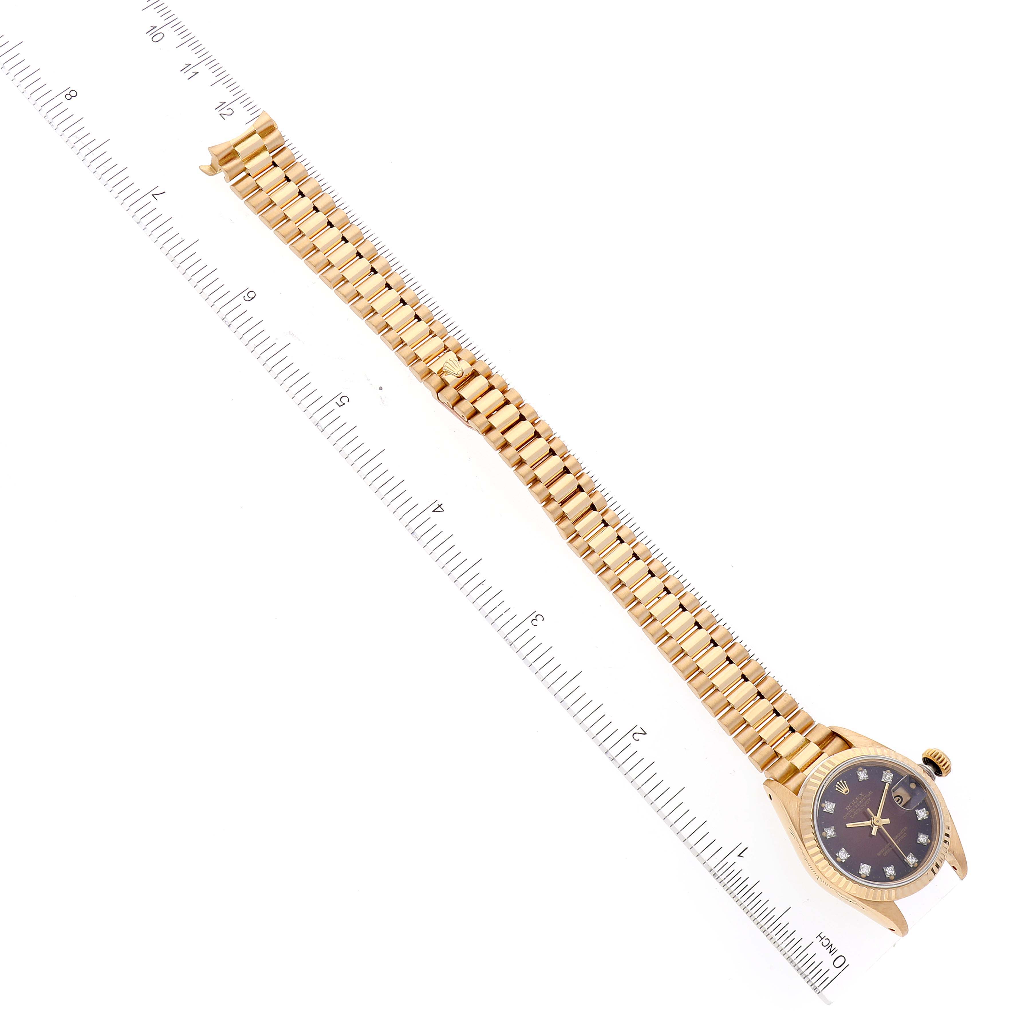The image shows a top-down view of a Rolex President 69178 Women's Yellow Gold Red Dial 69178 Women's Yellow Gold Red Dial watch, including its gold bracelet and diamond-studded face, aligned next to a ruler.