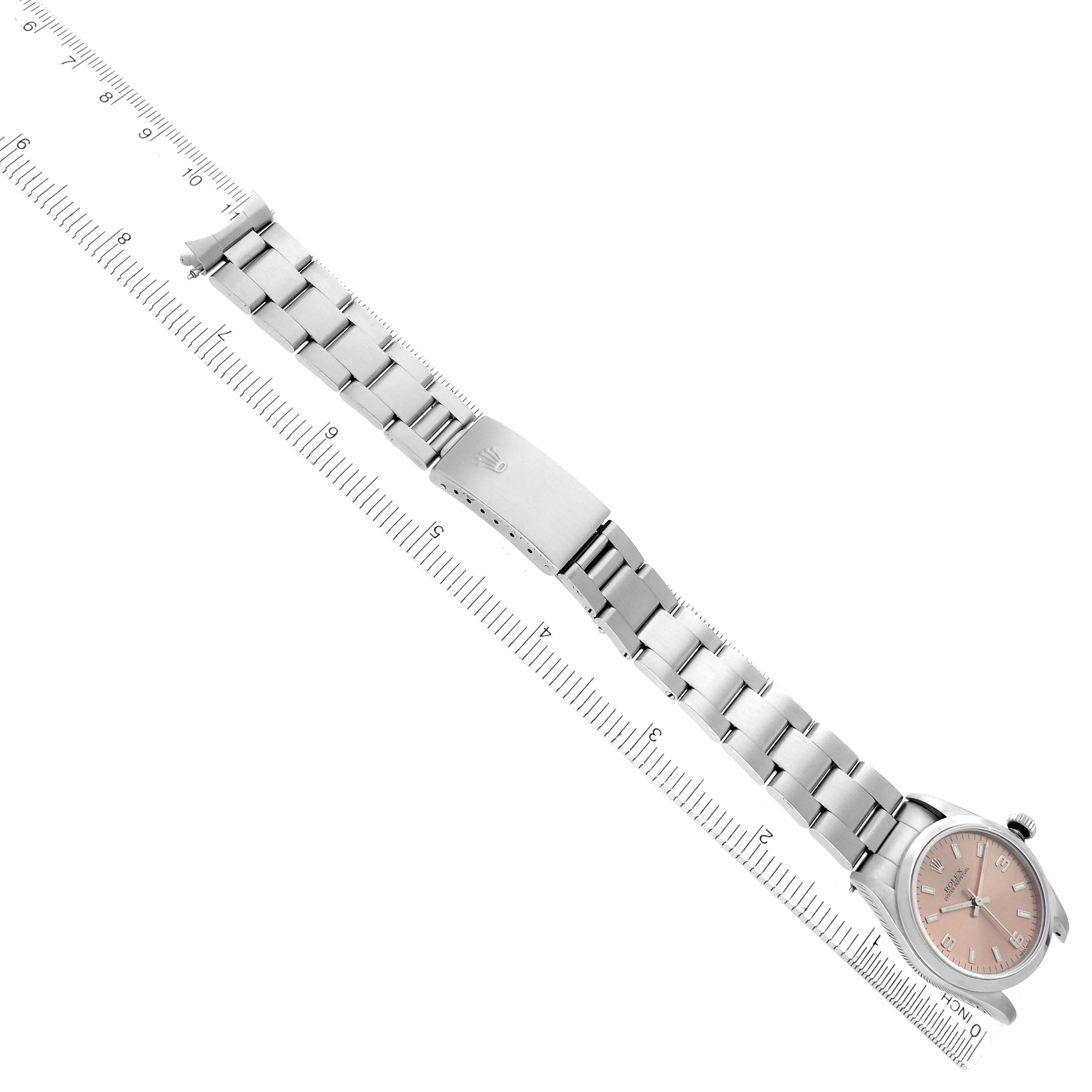 The Rolex Oyster Perpetual model 67480 is shown from a top-down angle, displaying its full metal bracelet, clasp, and face against a measuring ruler.