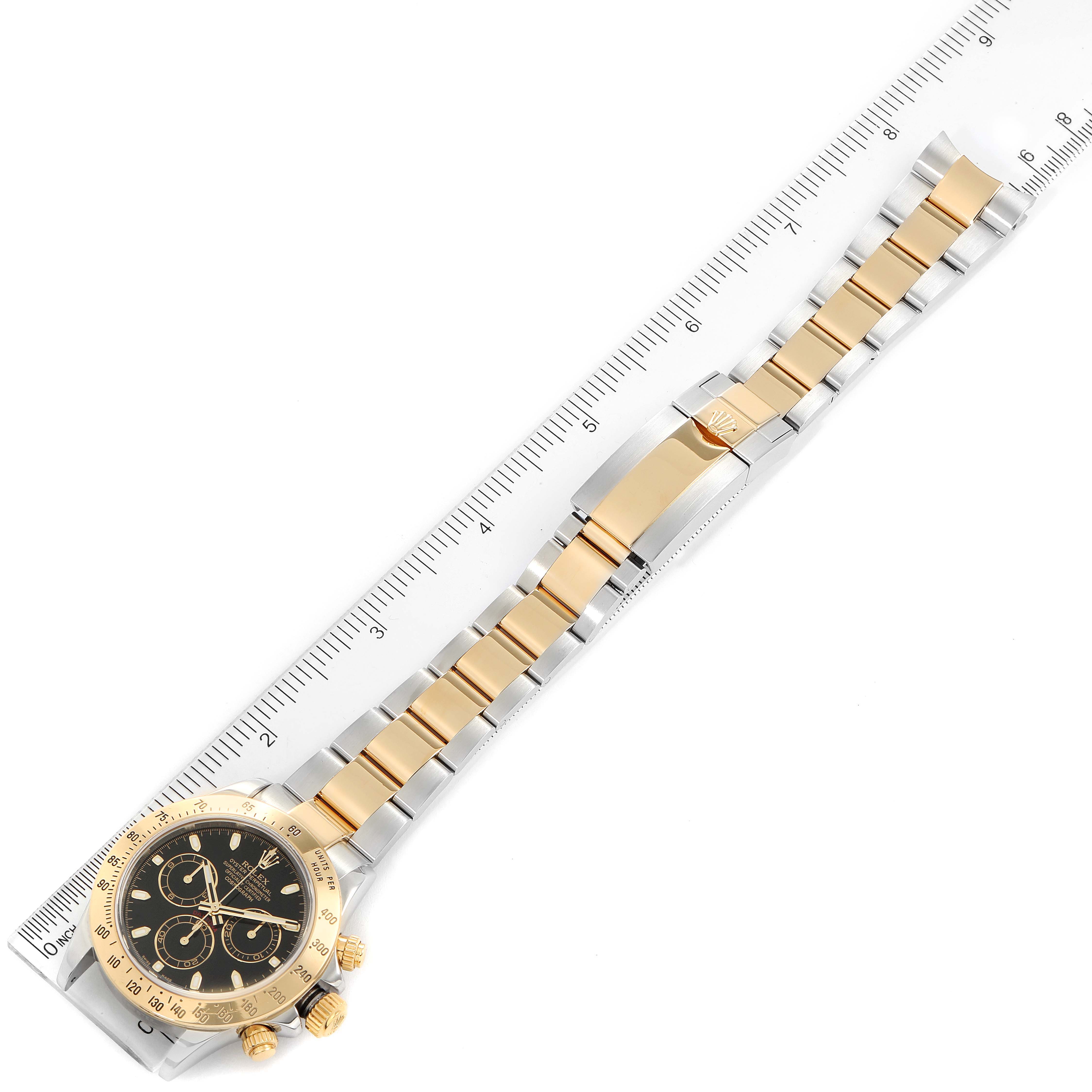 The image shows a Rolex Daytona 116523 Men's Steel and Gold (two tone) Black Dial 116523 Men's Steel and Gold (two tone) Black Dial watch laid flat against a ruler, showcasing its gold and silver bracelet and black dial face.