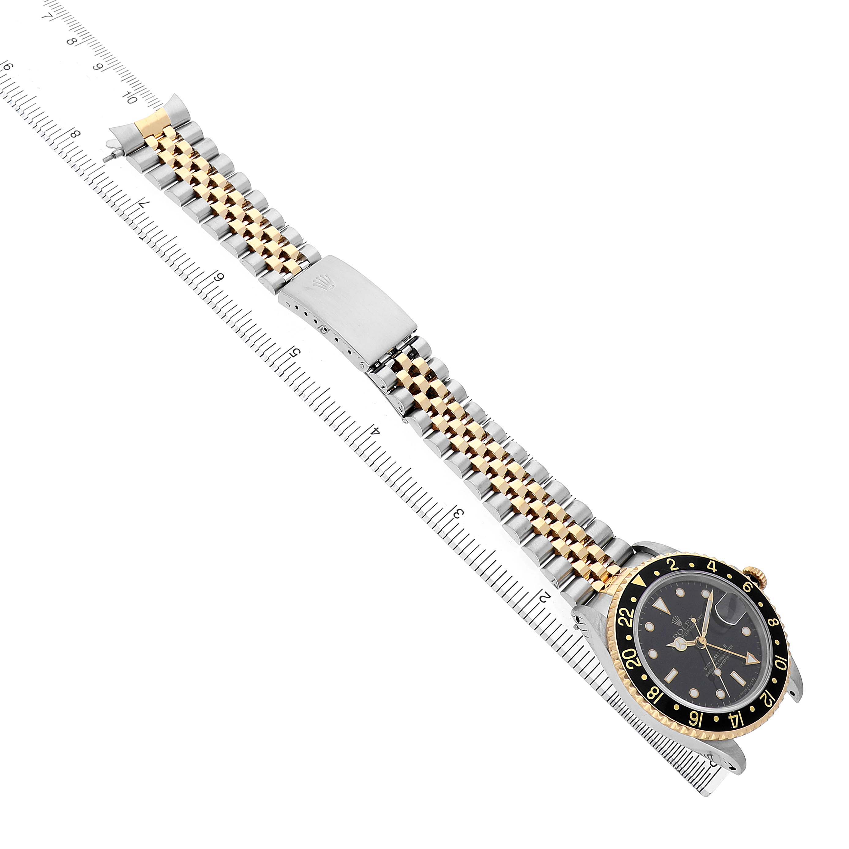 The Rolex GMT-Master 16713 Men's Steel and Gold (two tone) Black Dial watch is shown from a top-down angle, displaying the face, bezel, and two-tone bracelet with a measuring ruler.

