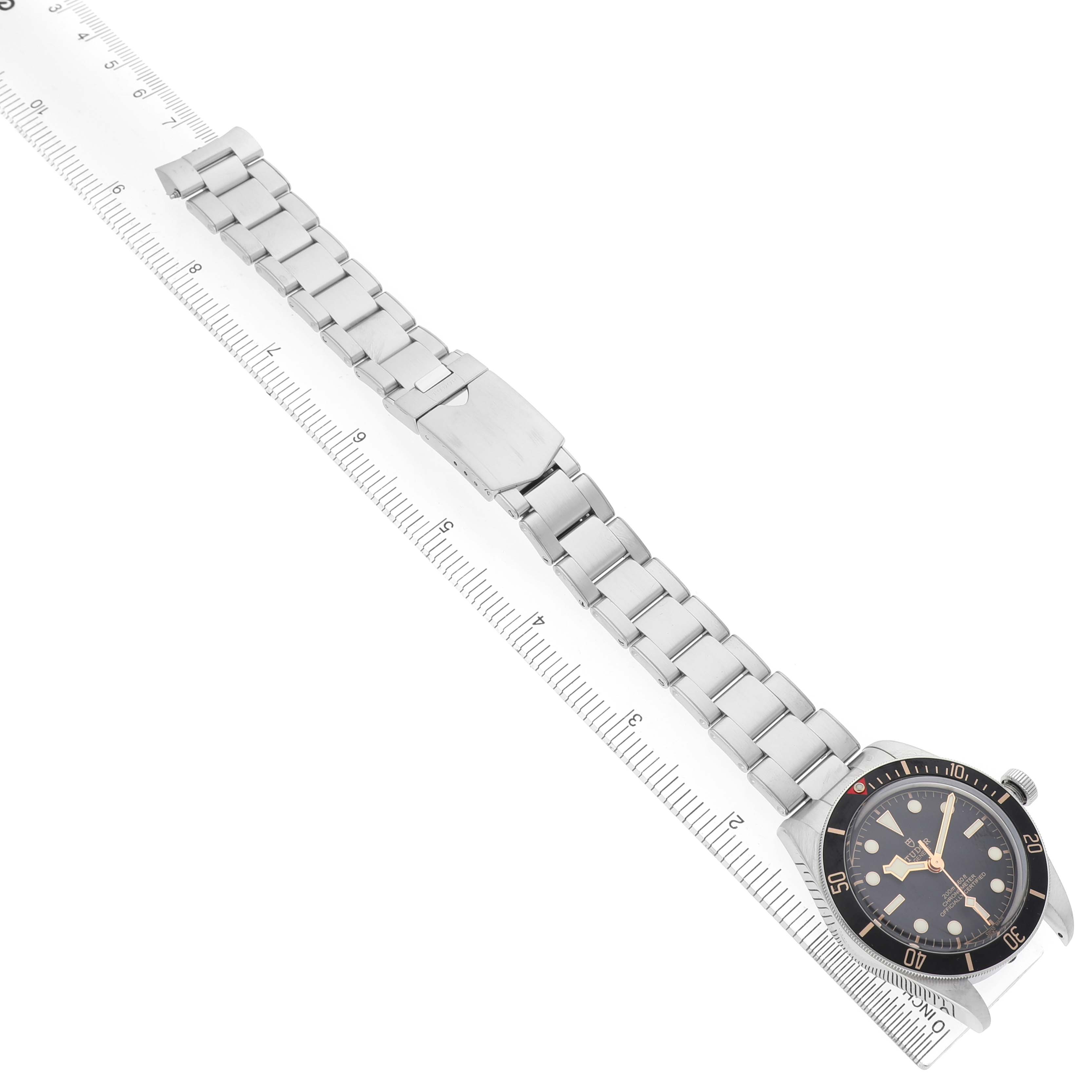 The Tudor Heritage Black Bay M79030N-0001 Men's Stainless Steel Black Dial watch is shown from an angled top view, displaying the full bracelet and clasp alongside a ruler.