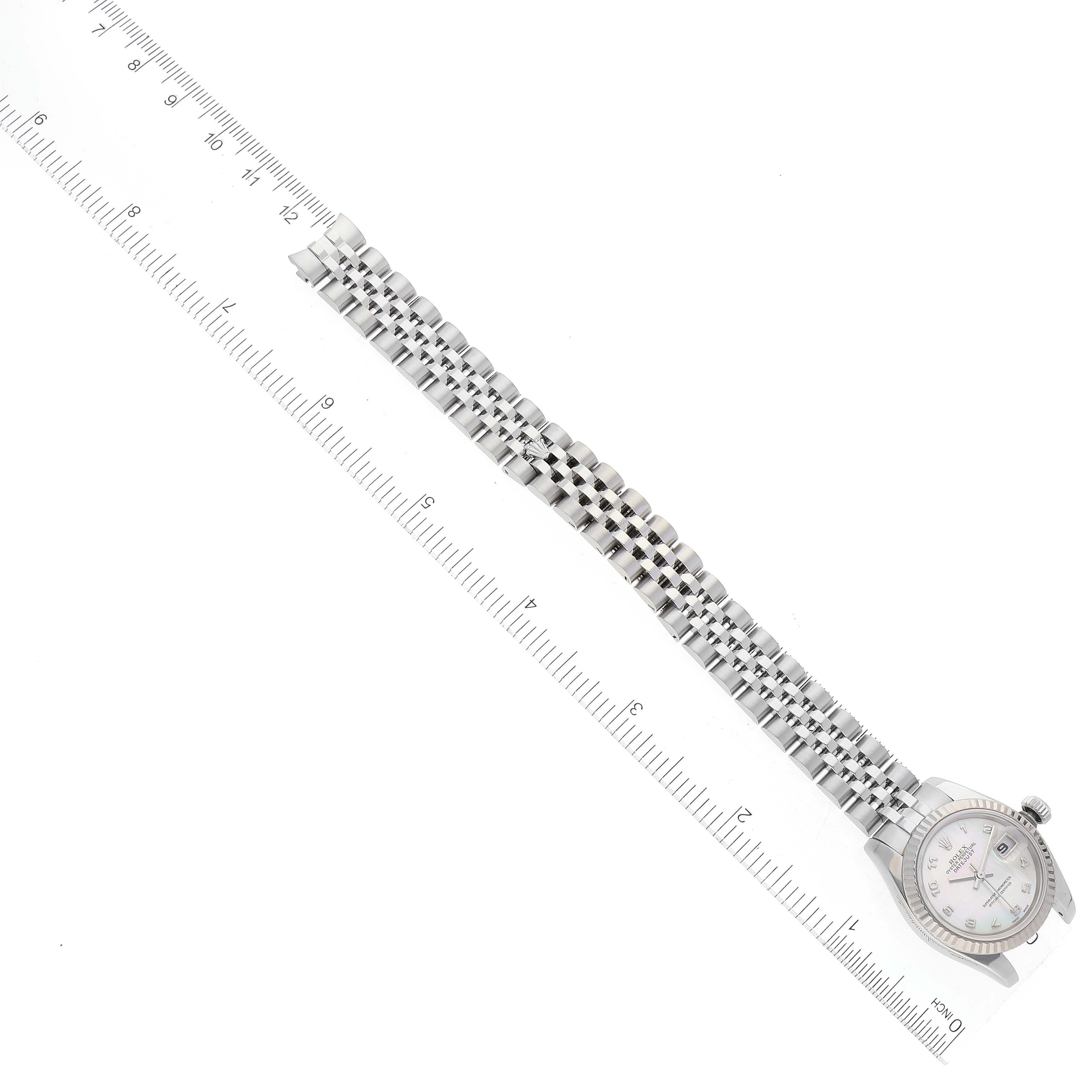 The image shows a Rolex Datejust 179174 Women
s Stainless Steel Mother of Pearl Dial 179174 Women
s Stainless Steel Mother of Pearl Dial watch from a top-down angle, highlighting the face, bezel, crown, and bracelet, alongside a ruler.