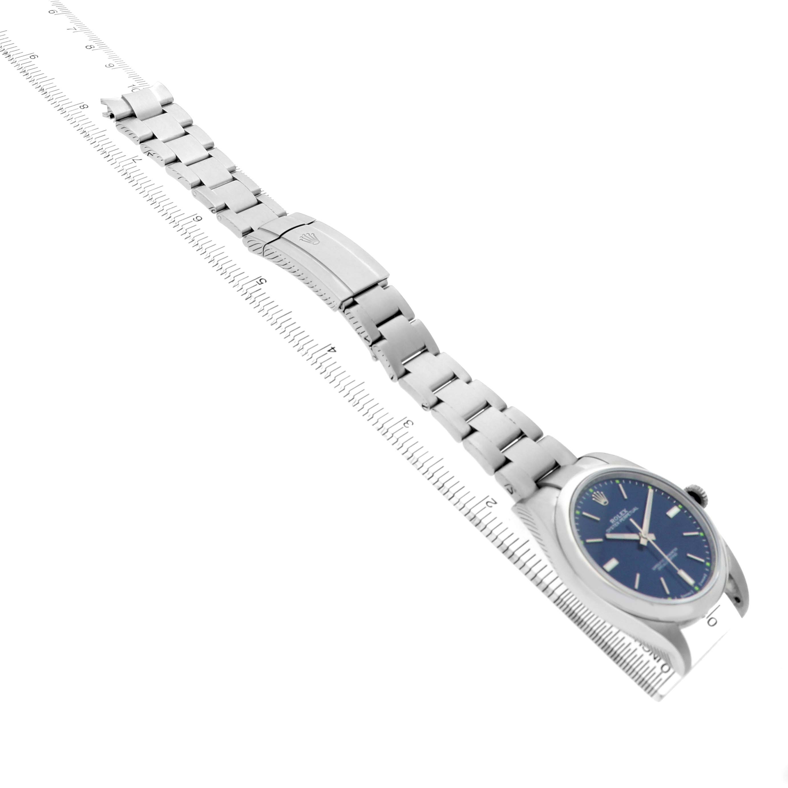 The Rolex Oyster Perpetual 114300 Men's Stainless Steel Blue Dial watch is shown at an angle highlighting the face, bracelet, and clasp aligned along a ruler.