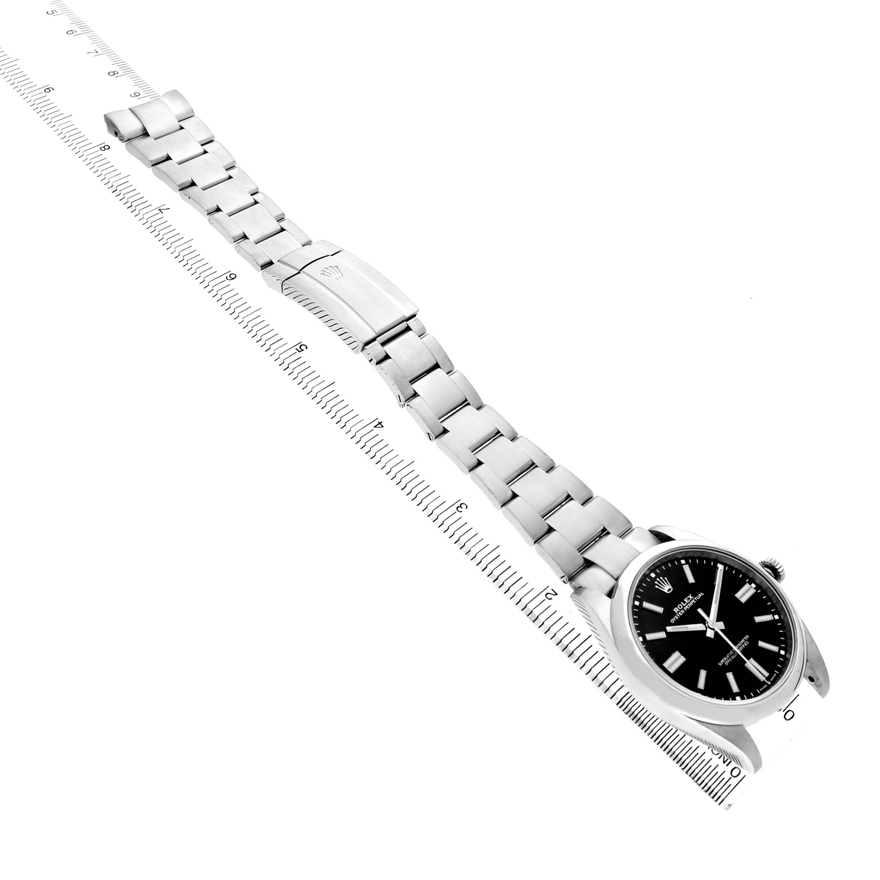 The image shows a Rolex Oyster Perpetual M124300-0002 Men's Stainless Steel Black Dial watch at an oblique angle, highlighting the face, bracelet, and clasp alongside a ruler.
