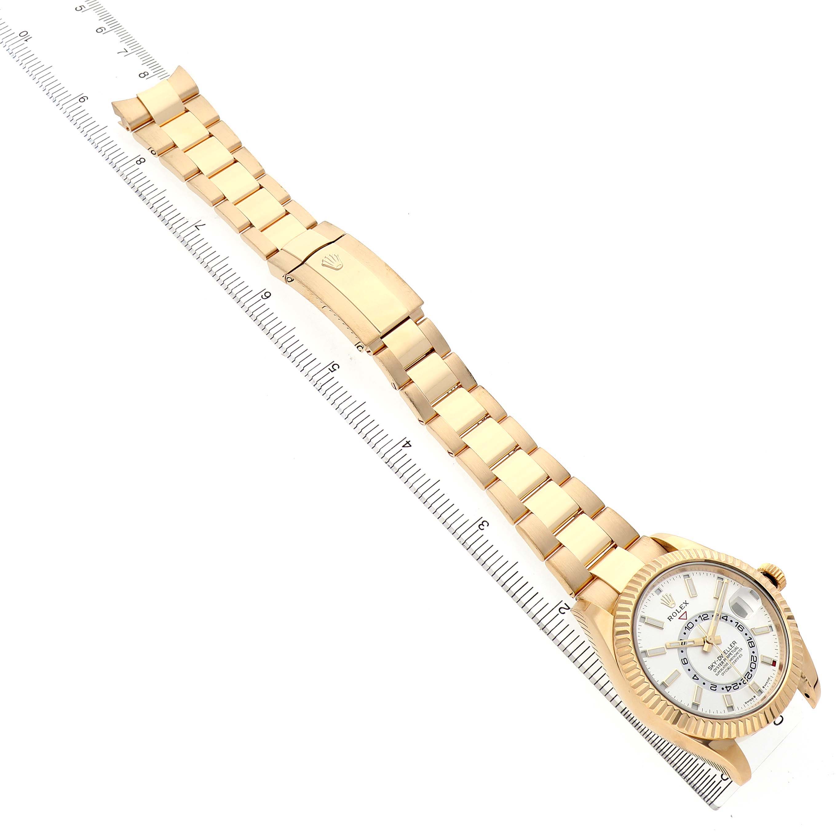 The Rolex Sky-Dweller 336938 Men's Yellow Gold White Dial watch is shown diagonally with its face, bracelet, and clasp visible alongside a ruler for scale.