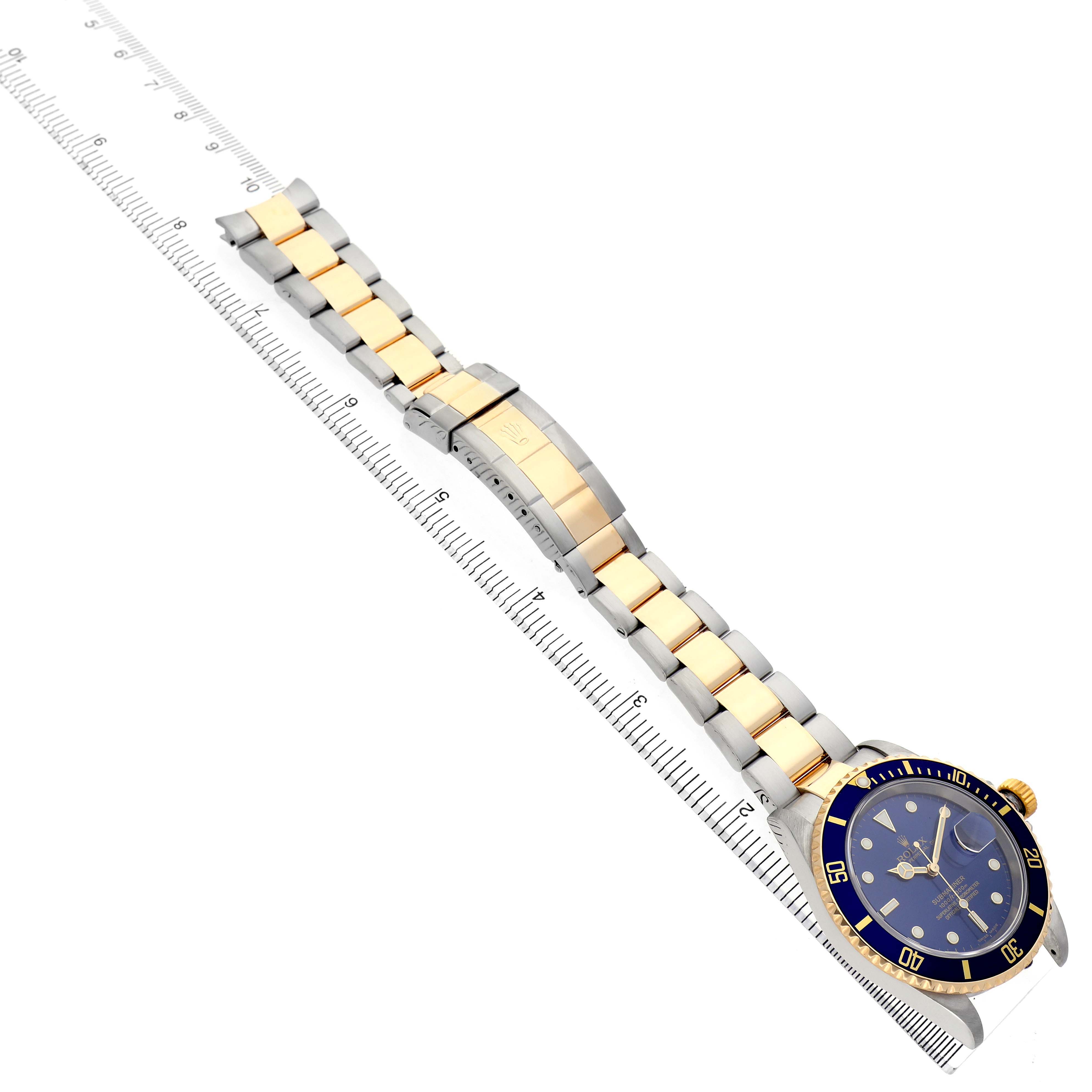 The Rolex Submariner 16613 Men's Steel and Gold (two tone) Blue Dial watch is angled diagonally, showing the face, bezel, crown, and full bracelet with a measuring ruler alongside.