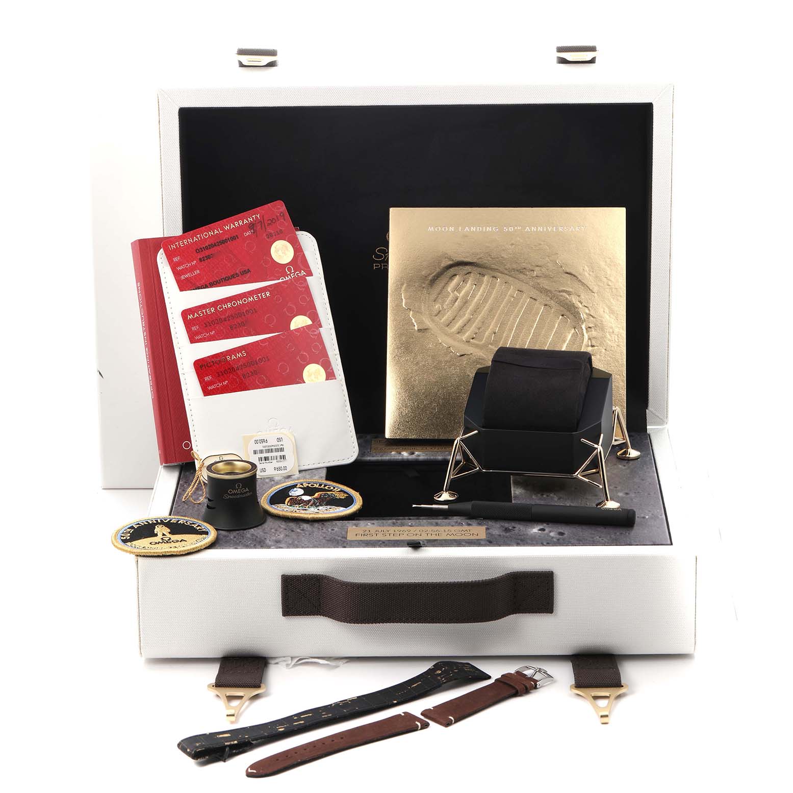 The image shows the Omega Speedmaster model 310.20.42.50.01.001 packaging, including watch accessories, patches, cards, and a lunar module display stand.