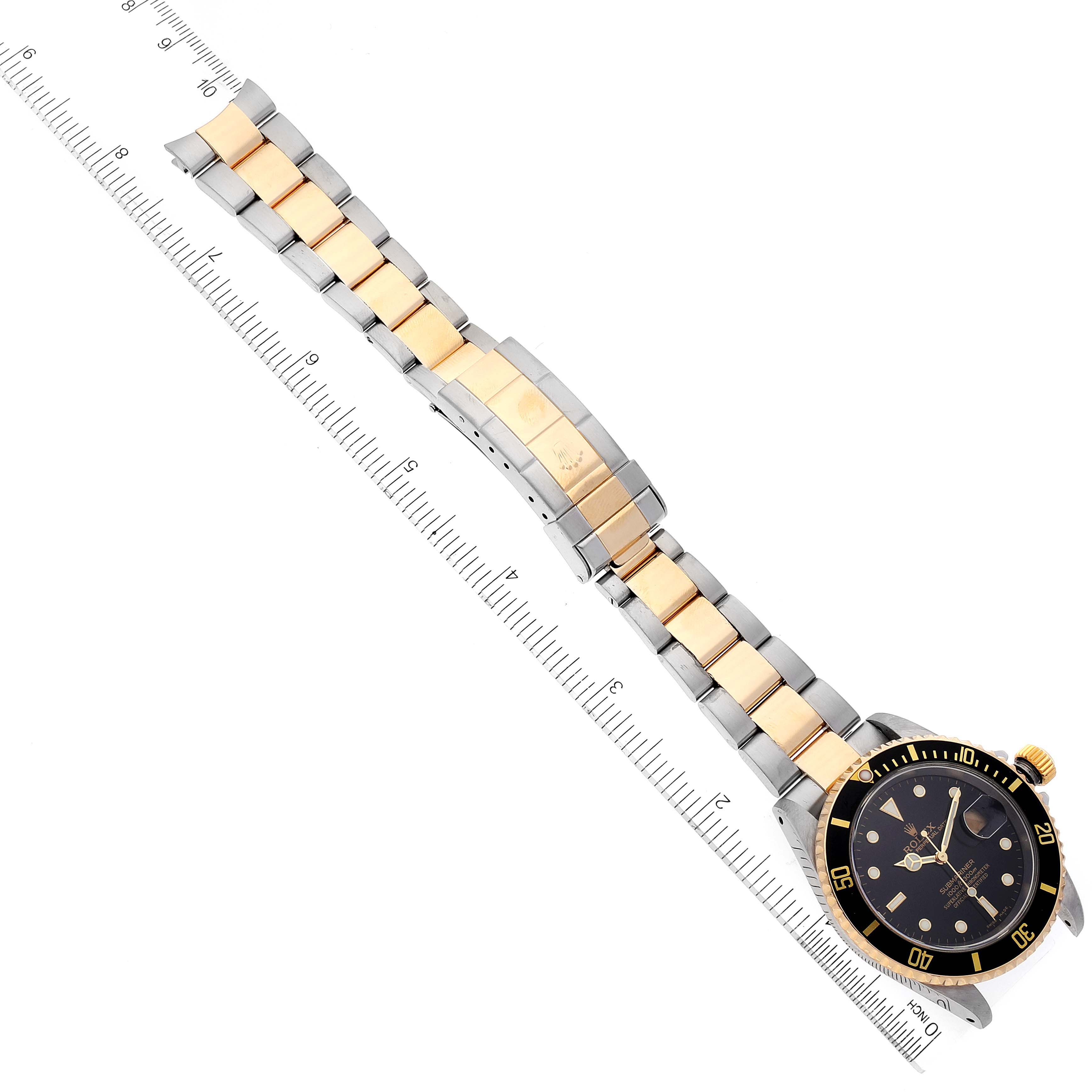 The image shows a Rolex Submariner 16613 Men's Steel and Gold (two tone) Black Dial 16613 Men's Steel and Gold (two tone) Black Dial watch, including its gold and silver bracelet, flat on a ruler for scale.