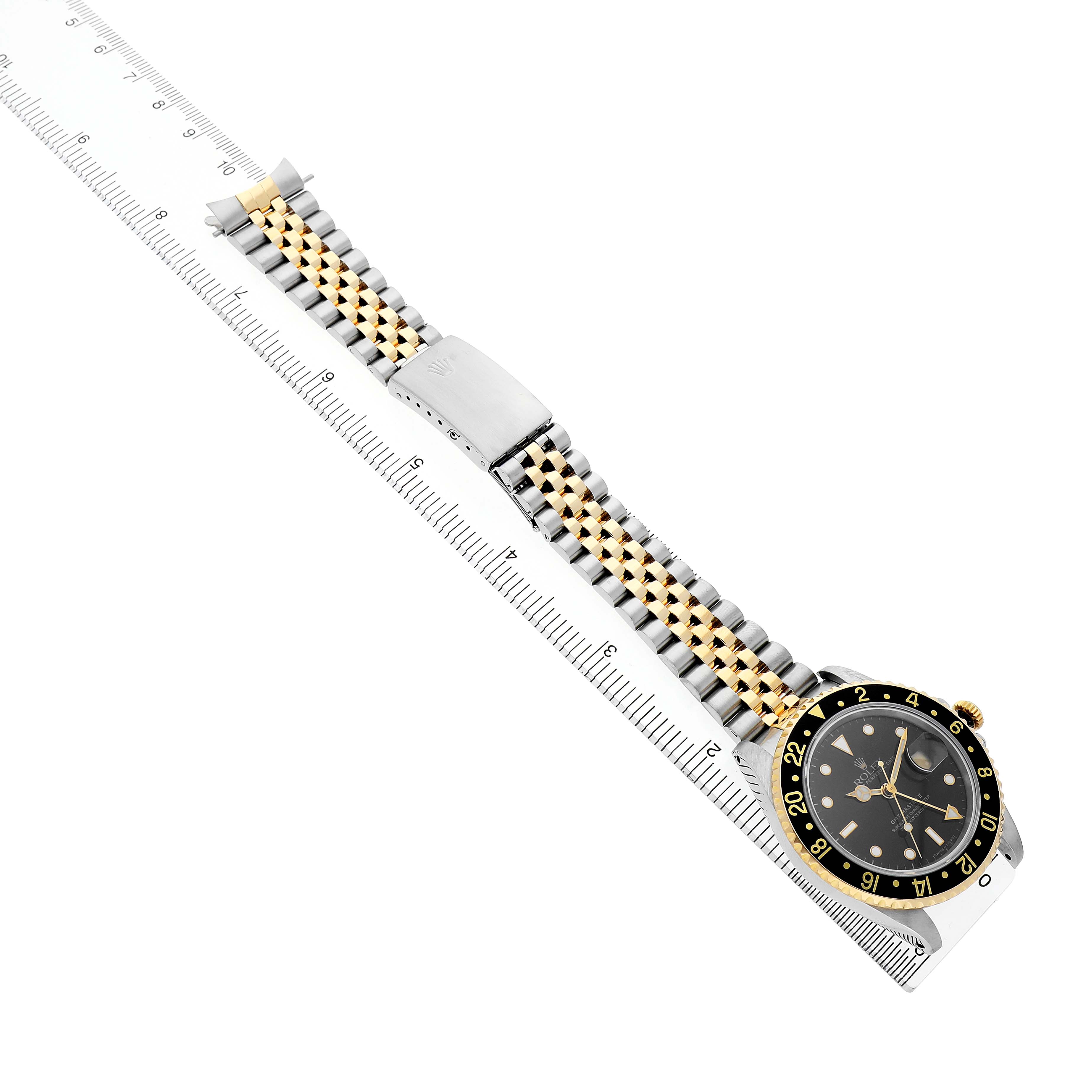 The Rolex GMT-Master 16713 Men's Steel and Gold (two tone) Black Dial watch is shown from an angled top view, displaying the face, bezel, and bracelet laid flat alongside a ruler.