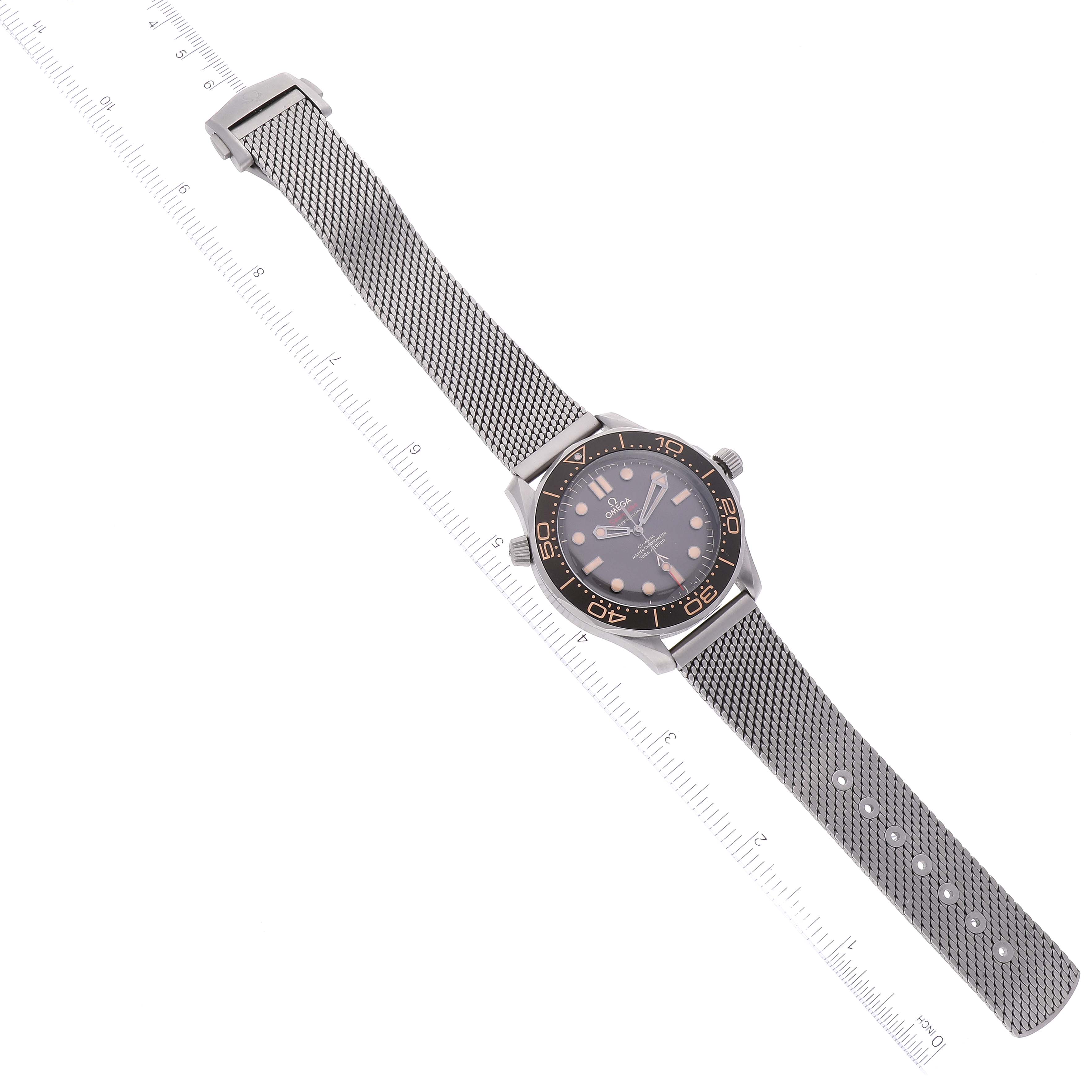 The image shows an Omega Seamaster 210.90.42.20.01.001 Men's Titanium Brown Dial 210.90.42.20.01.001 Men's Titanium Brown Dial watch laid flat, with a mesh strap and markings visible on a ruler beneath it.