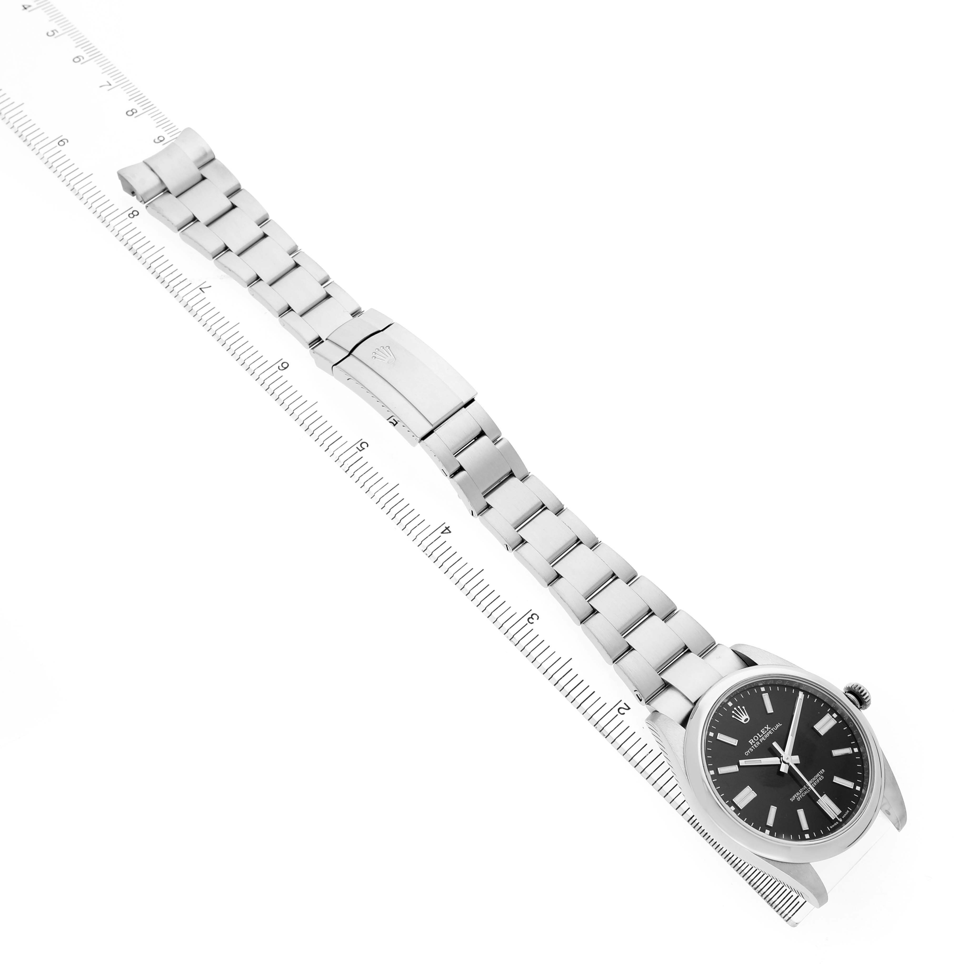 The image shows a Rolex Oyster Perpetual M124300-0002 Men's Stainless Steel Black Dial watch from a top-down angle, highlighting the bracelet, clasp, and watch face alongside a ruler.