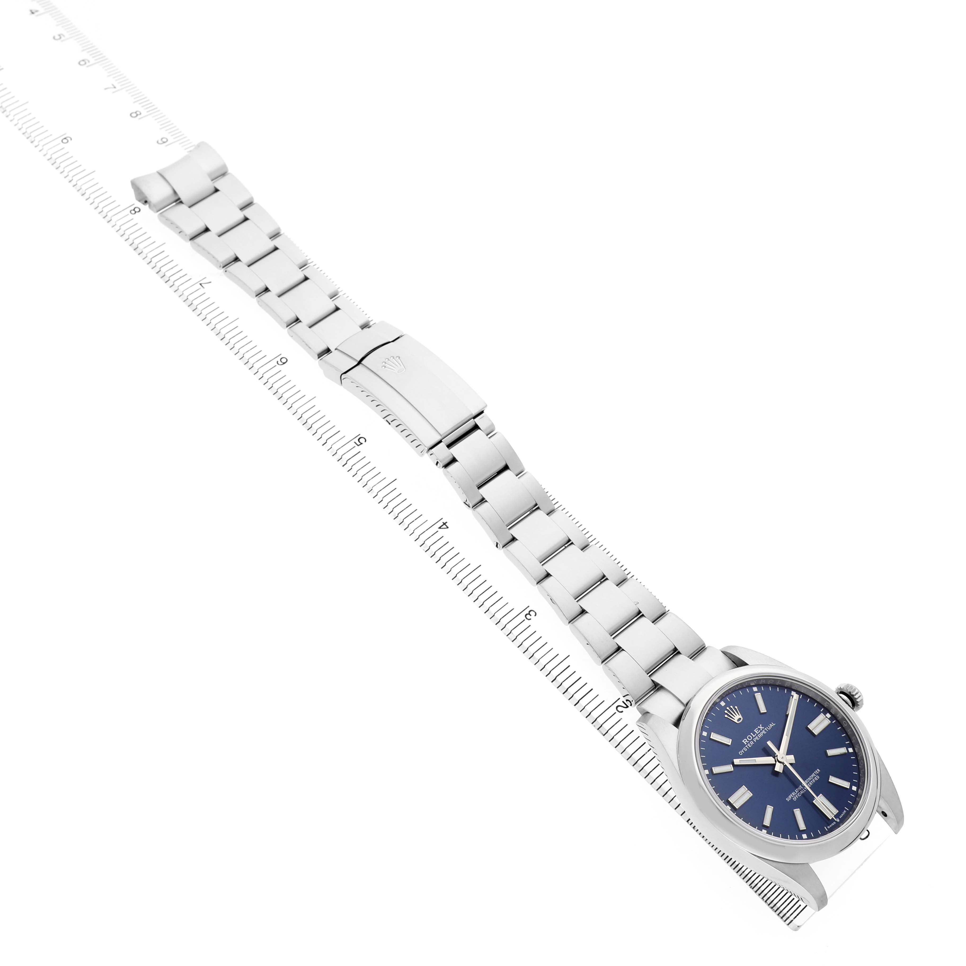 The image shows a Rolex Oyster Perpetual 124300 Men's Stainless Steel Blue Dial watch at an angle, highlighting its blue dial and metal bracelet laid on a ruler.