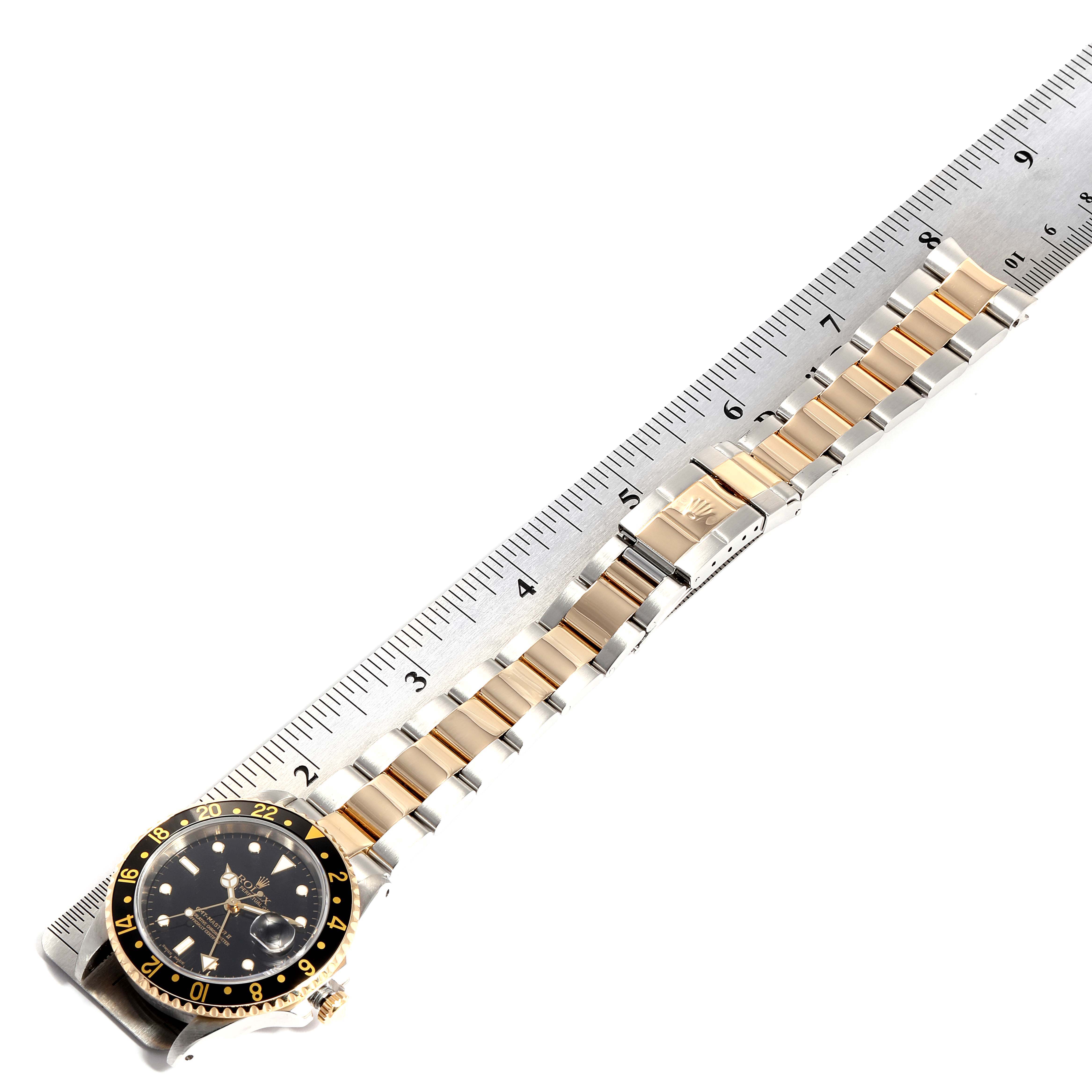 The image shows a Rolex GMT-Master 16713 Men
s Steel and Gold (two tone) Black Dial 16713 Men
s Steel and Gold (two tone) Black Dial watch laid flat next to a ruler, displaying the face, bezel, and bracelet.