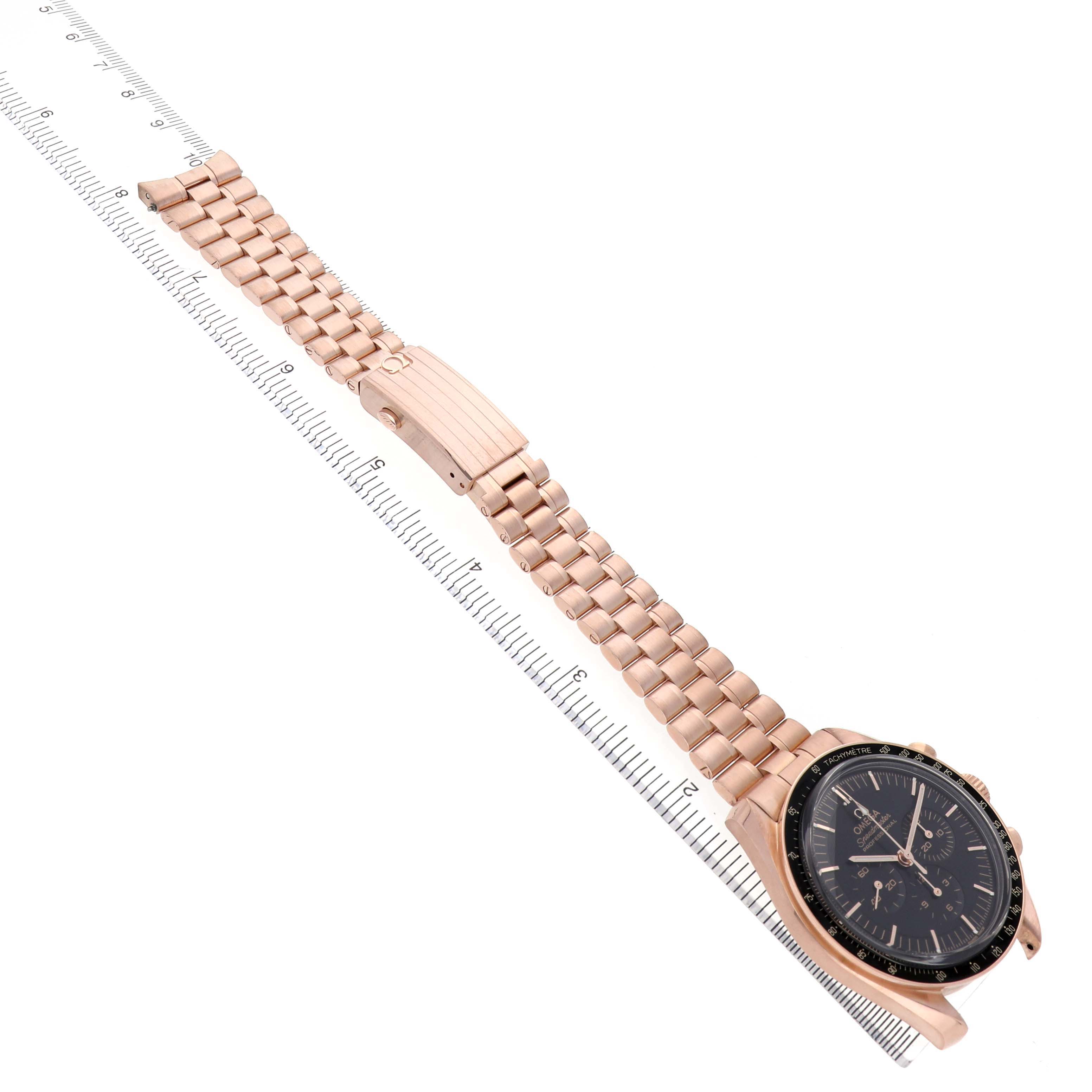 The Omega Speedmaster 310.60.42.50.01.001 Men's Rose Gold Black Dial watch is shown at a diagonal angle, displaying the face, bezel, and full metal bracelet alongside a ruler.