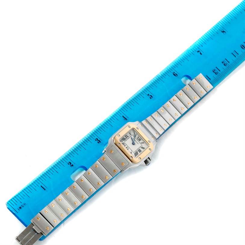 The image shows a Santos model watch by Cartier with its face and full bracelet laid next to a blue ruler for scale.