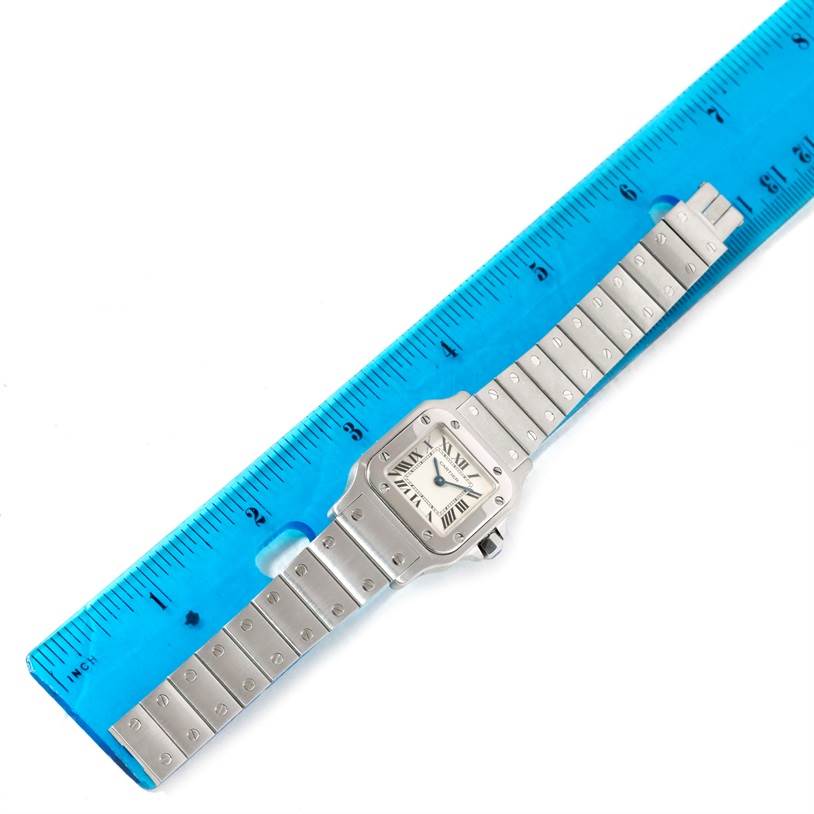 The image shows a full view of a Santos model Cartier watch, lying adjacent to a blue ruler.